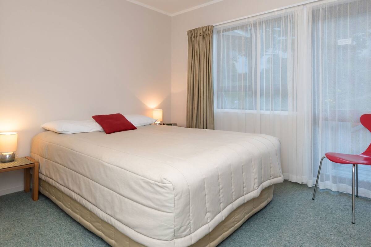 A hotel room with a bed, bedside lamps, a chair, and a window with sheer curtains.