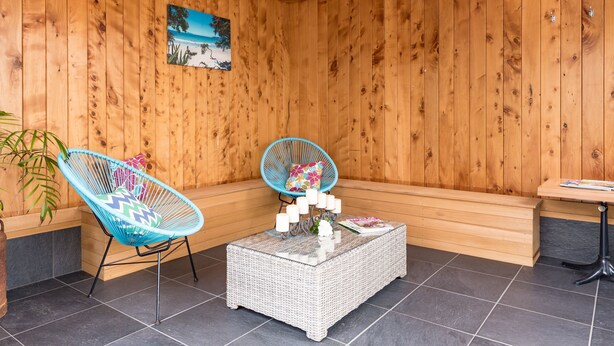 Een kamer met houten lambrisering, twee blauwe stoelen, een witte rieten tafel en een bank.