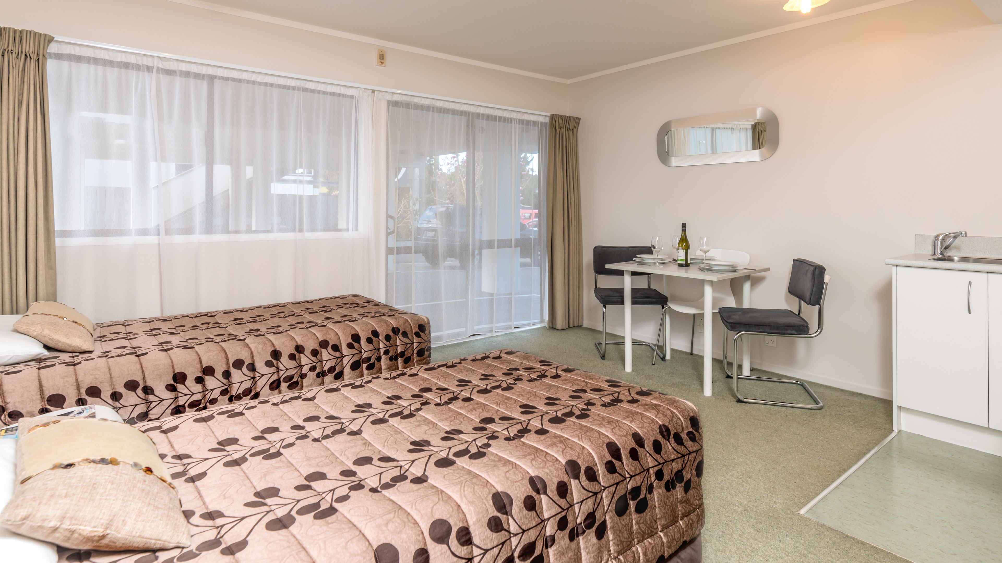 Habitación de hotel con dos camas, una mesa de comedor con sillas, una pequeña cocina y un amplio ventanal con cortinas.