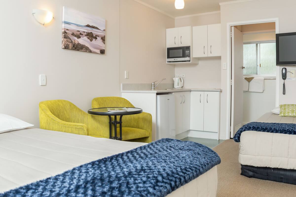 A hotel room with a bed, two yellow armchairs, a small table, a kitchenette with a microwave, and a TV.