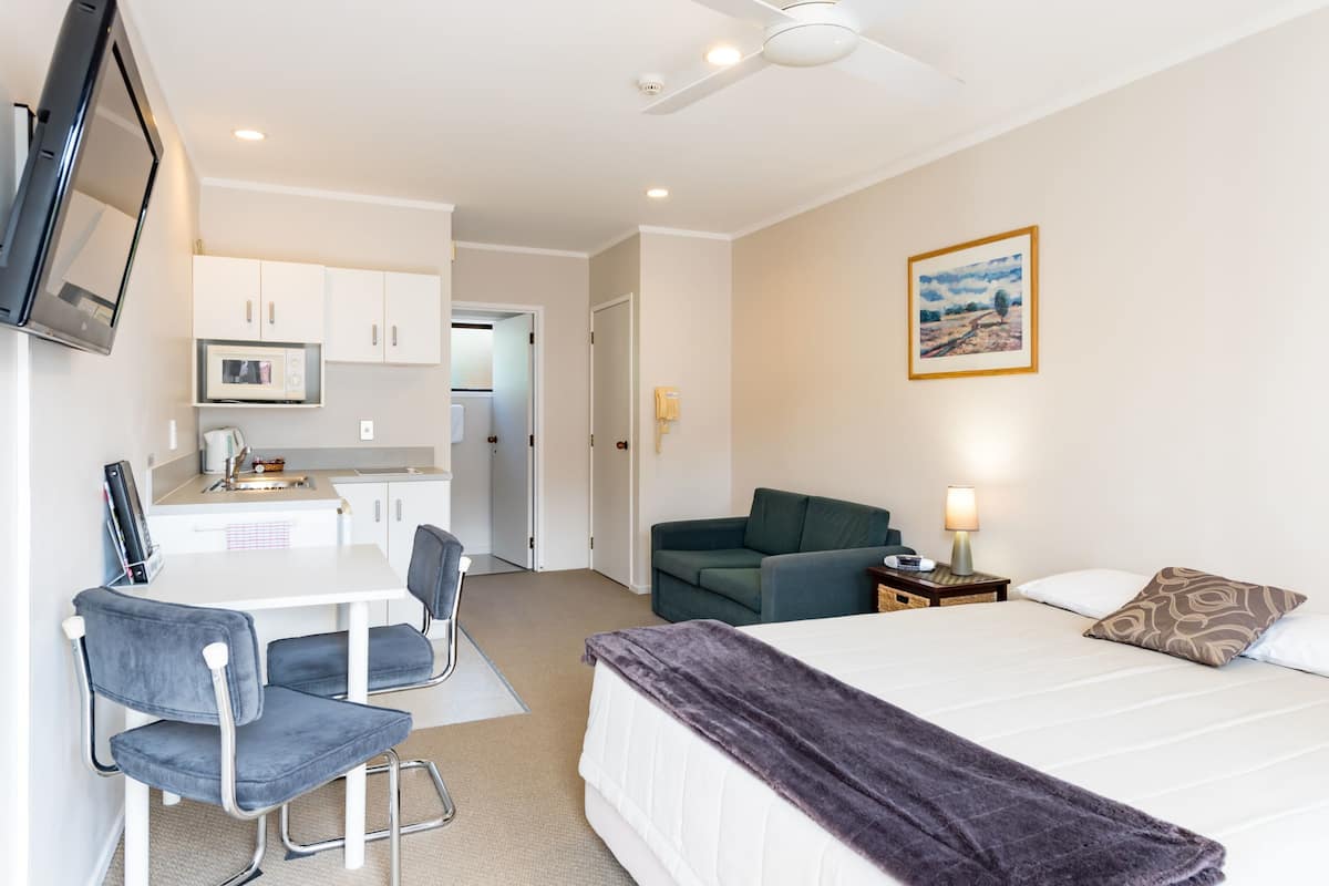 A hotel room with a bed, a desk with a chair, a kitchenette, a ceiling fan, and a painting on the wall.