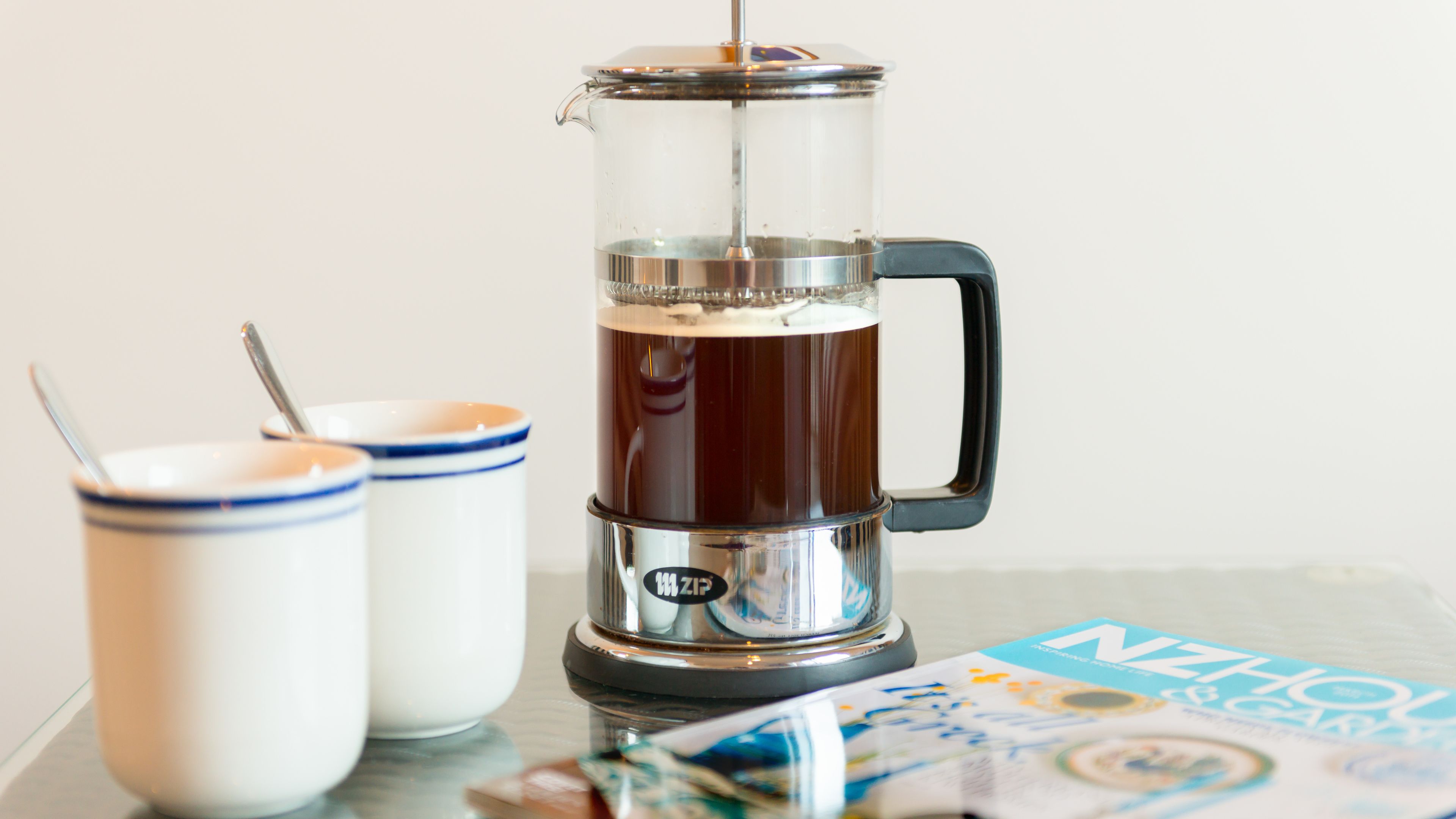 A coffee maker with a clear glass carafe and a metal handle, two white mugs with blue trim, and a magazine on a table.