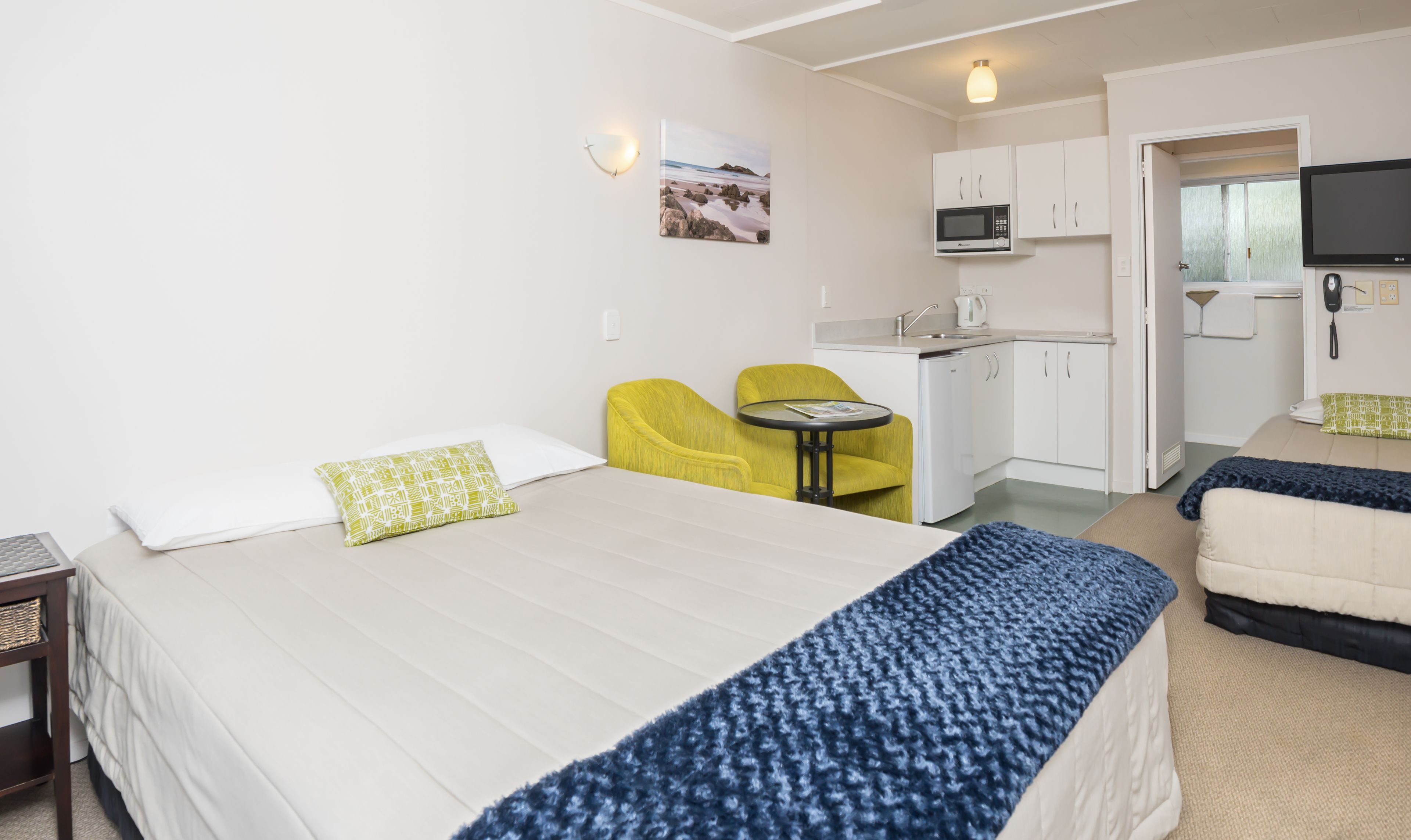 A hotel room with a bed, a small desk, a chair, a kitchenette, and a TV.