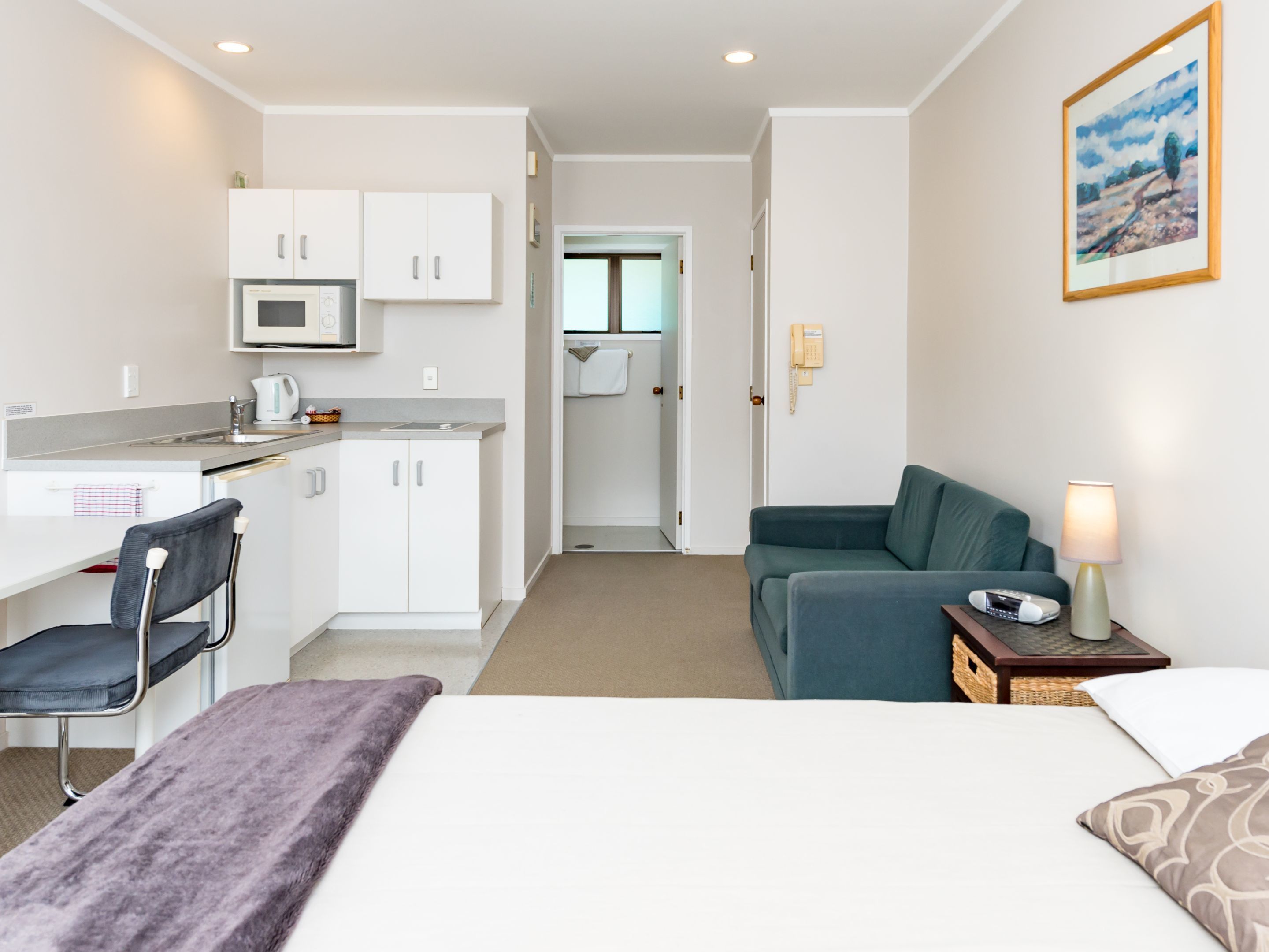 A hotel room with a bed, a desk with a chair, a kitchenette, a sofa, and a bedside table with a lamp.