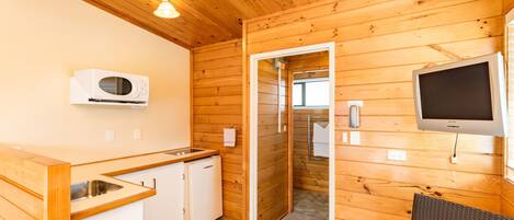 A compact kitchen with wooden walls, a built-in microwave, a small refrigerator, and a flat-screen TV mounted on the wall.