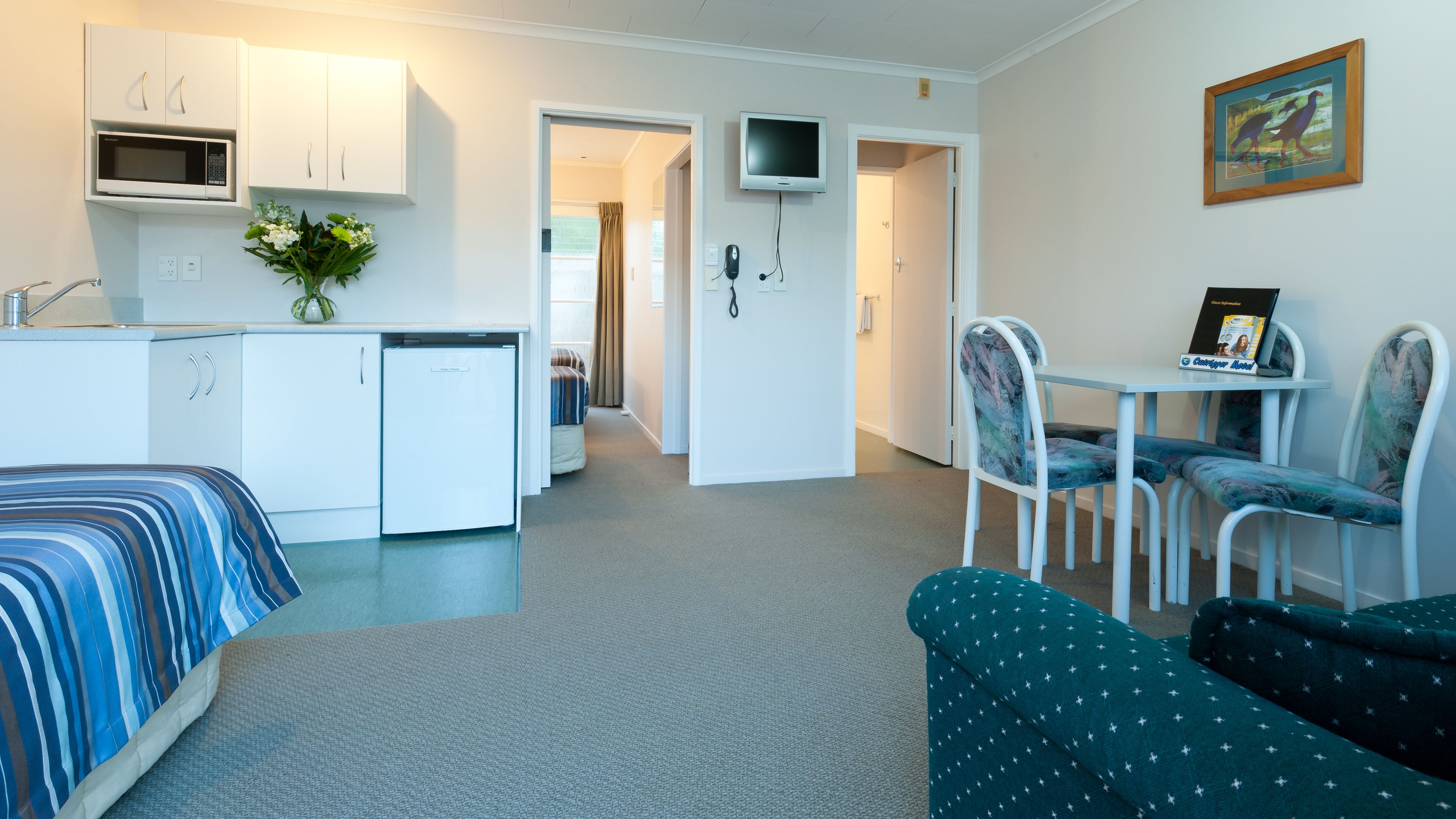 Habitación de hotel compacta con kitchenette, zona de comedor con mesa y sillas, cama con ropa de cama a rayas y televisor montado en la pared.