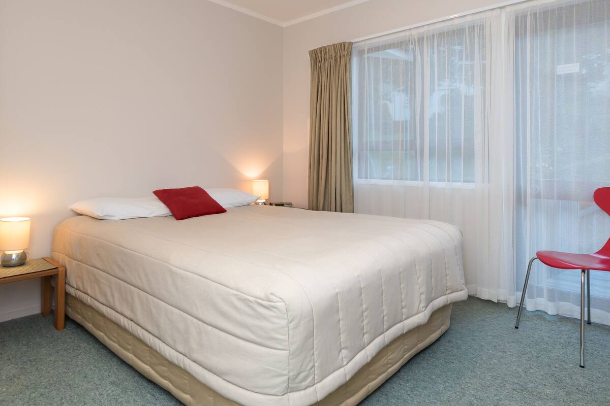 A neatly made bed with a red pillow, a bedside table with a lamp, and a red chair.