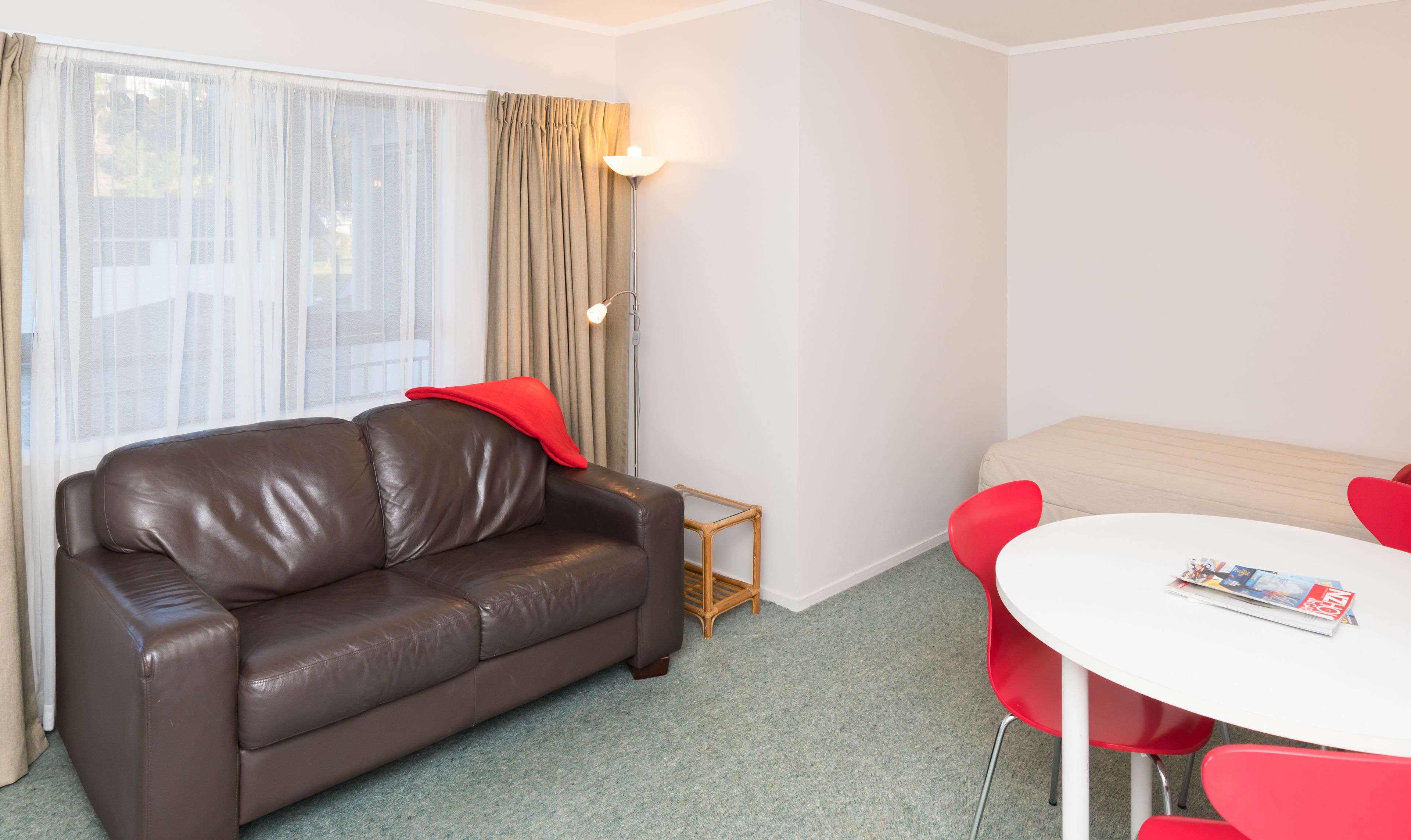 A modern living room with a brown leather sofa, a small wooden side table, a white round table with red chairs, and a window with sheer curtains.