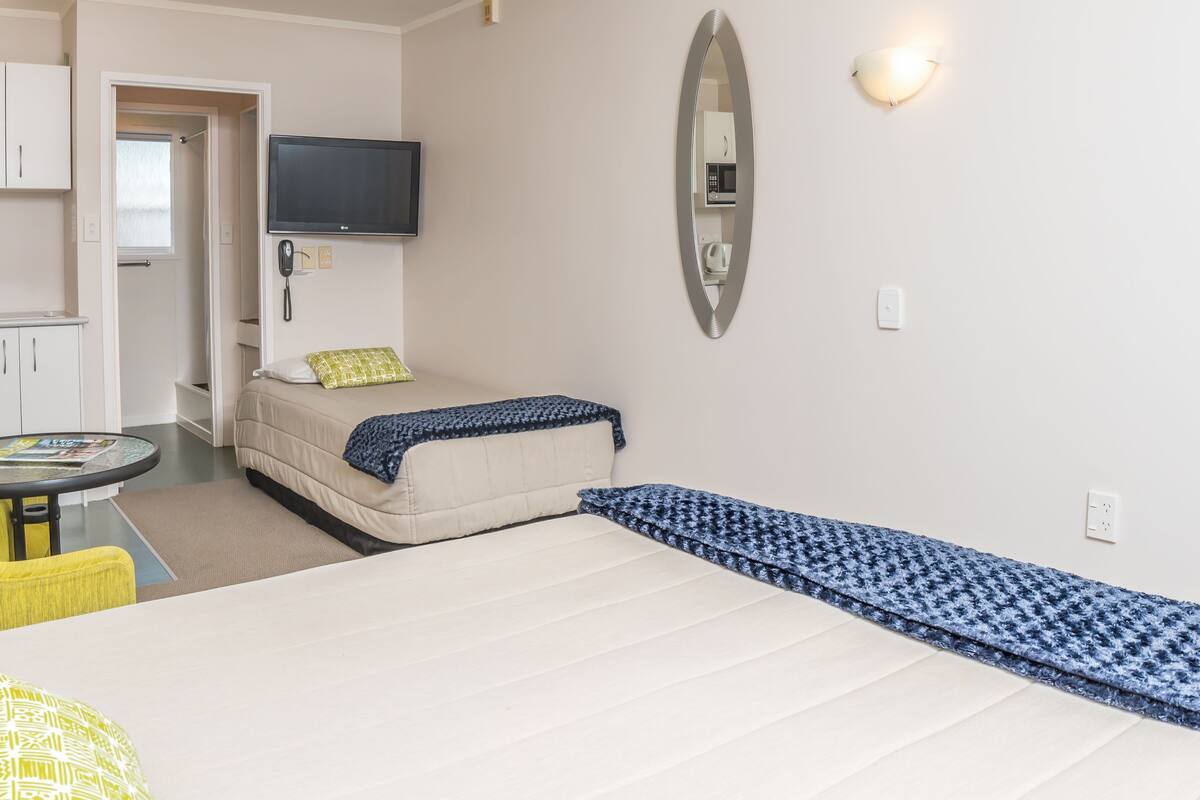 A hotel room with a bed, a small table, a yellow armchair, a kitchenette, and a TV.