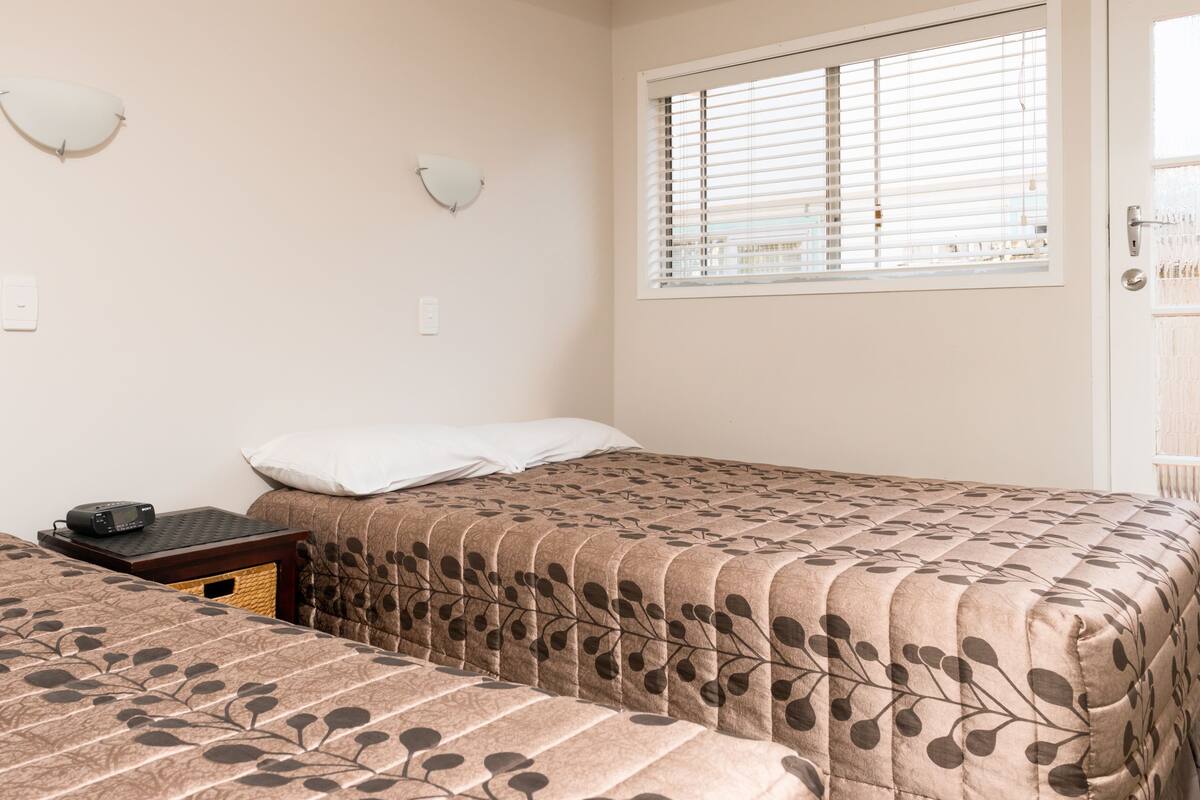A room with two beds, a bedside table with a phone, and a window with blinds.
