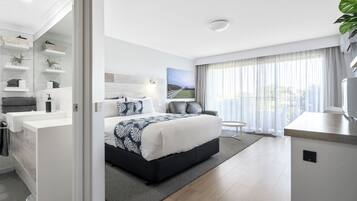 A modern hotel room with a large bed, a seating area, a bathroom with a vanity, and a view of the outdoors through the window.