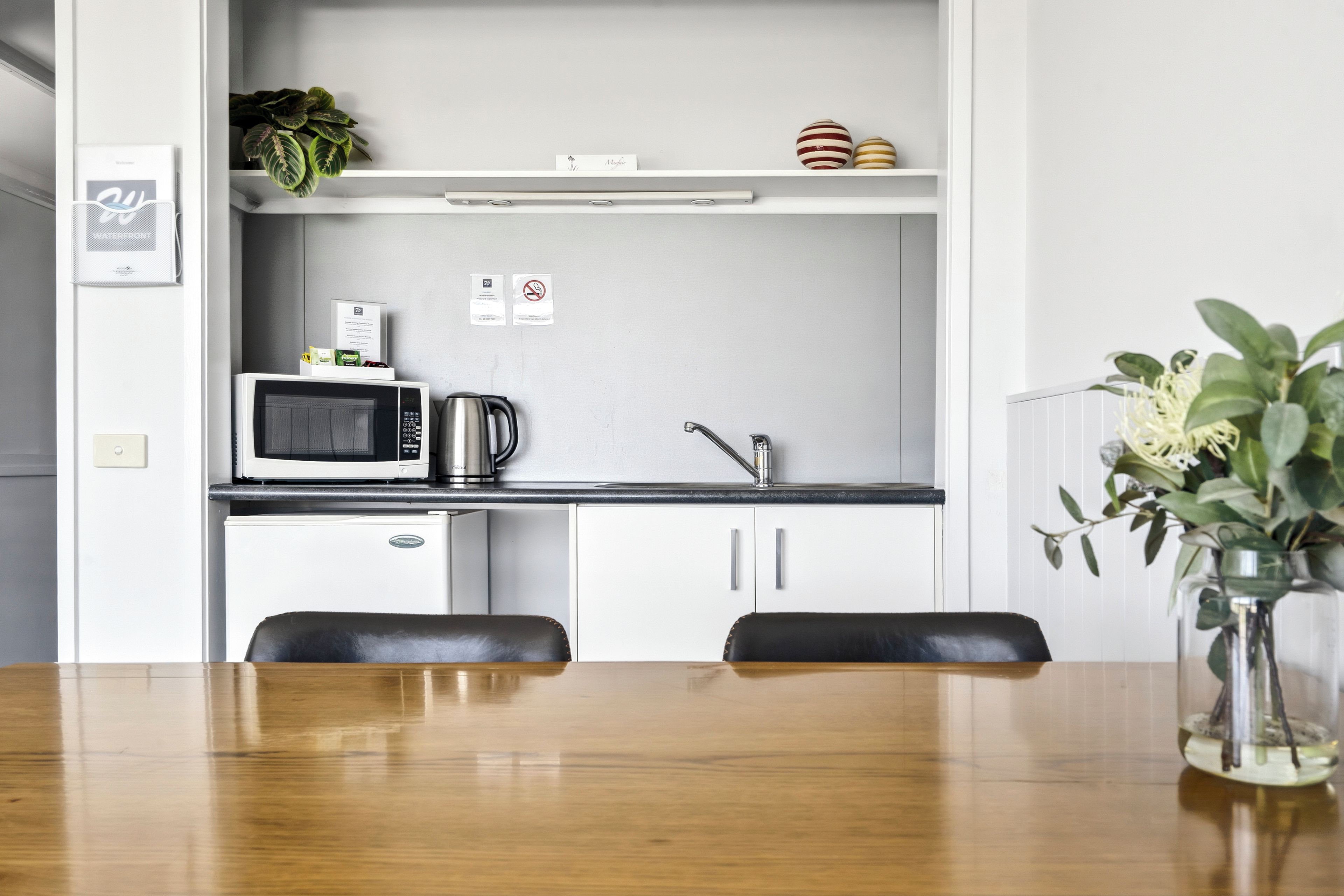 A compact kitchen with a microwave, kettle, and a coffee machine. There is a small refrigerator, a sink, and a cabinet above the counter. A dining table with chairs is in the foreground.
