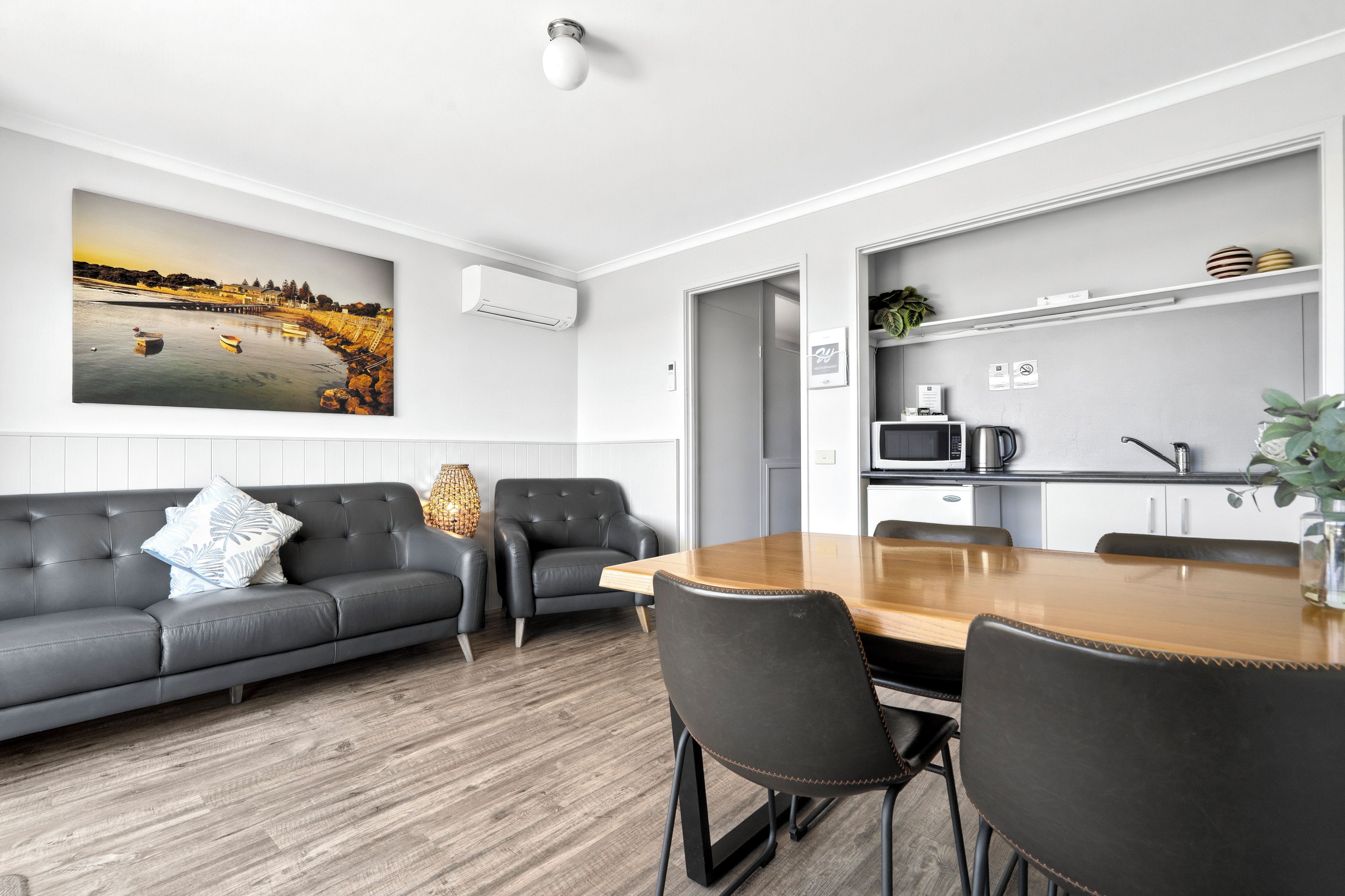 A modern living room with a grey sofa, a wooden dining table, and a kitchen area with a microwave and kettle.