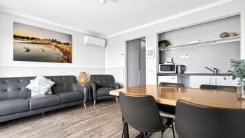 A modern living room with a grey sofa, a wooden dining table, and a kitchen area with a microwave and kettle.
