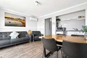 Un salon moderne avec un canapé gris, une table à manger en bois et un coin cuisine équipé d’un four à micro-ondes et d’une bouilloire.