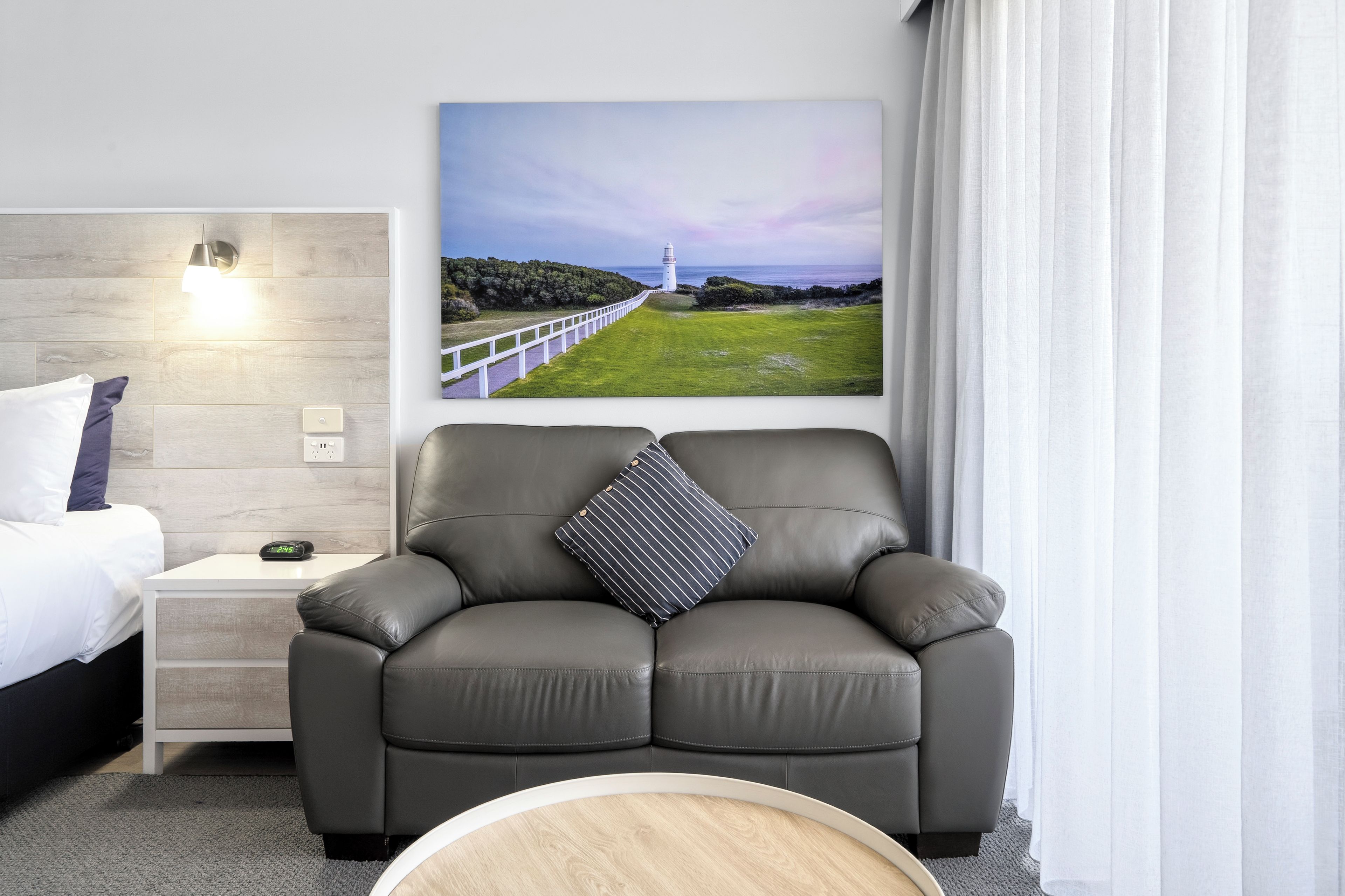 A modern hotel room with a bed, a leather armchair, a bedside table with a clock, and a painting of a coastal landscape.