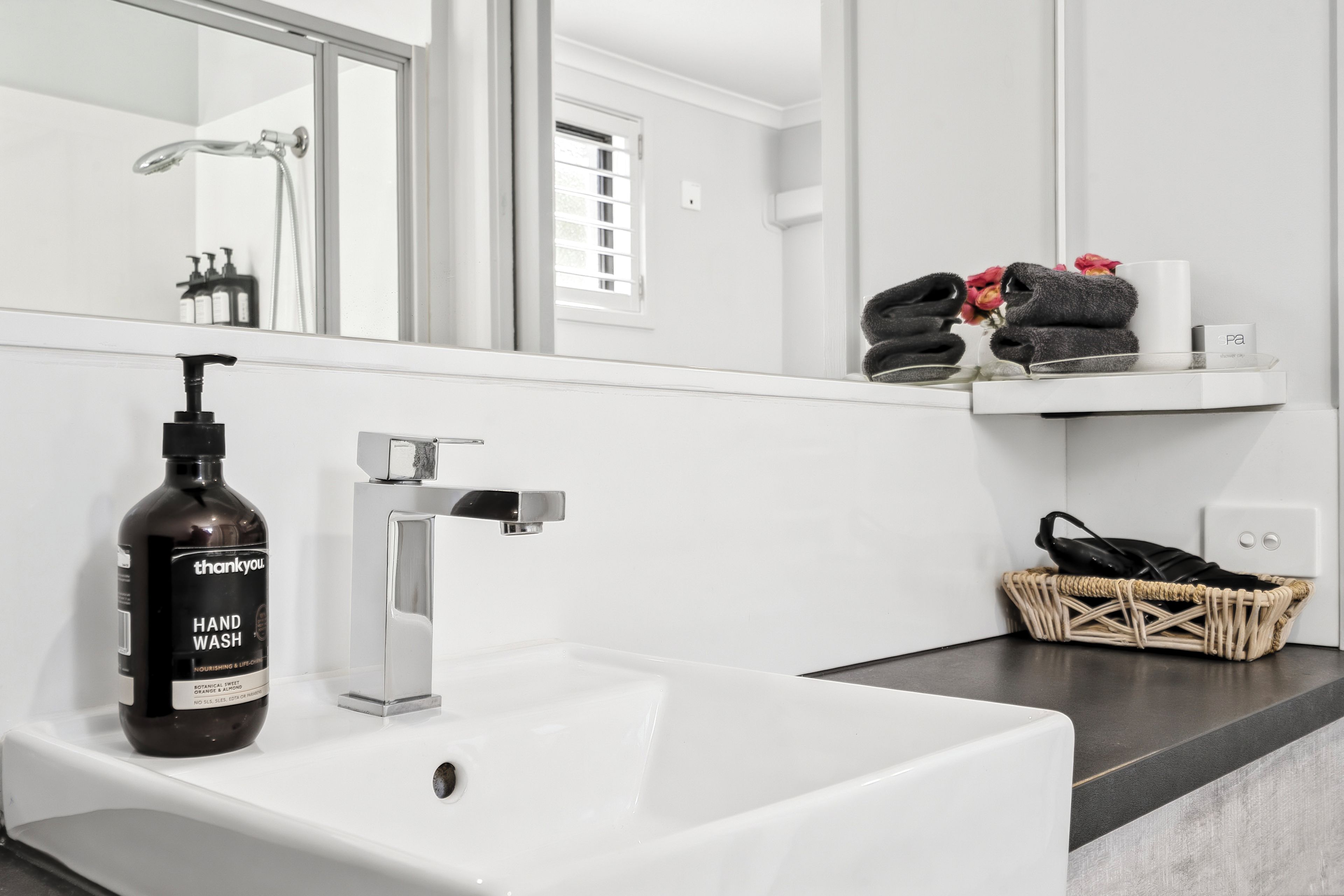 A bathroom with a white sink, a hand wash bottle, a modern faucet, a mirror, a window, and a towel rack with towels.