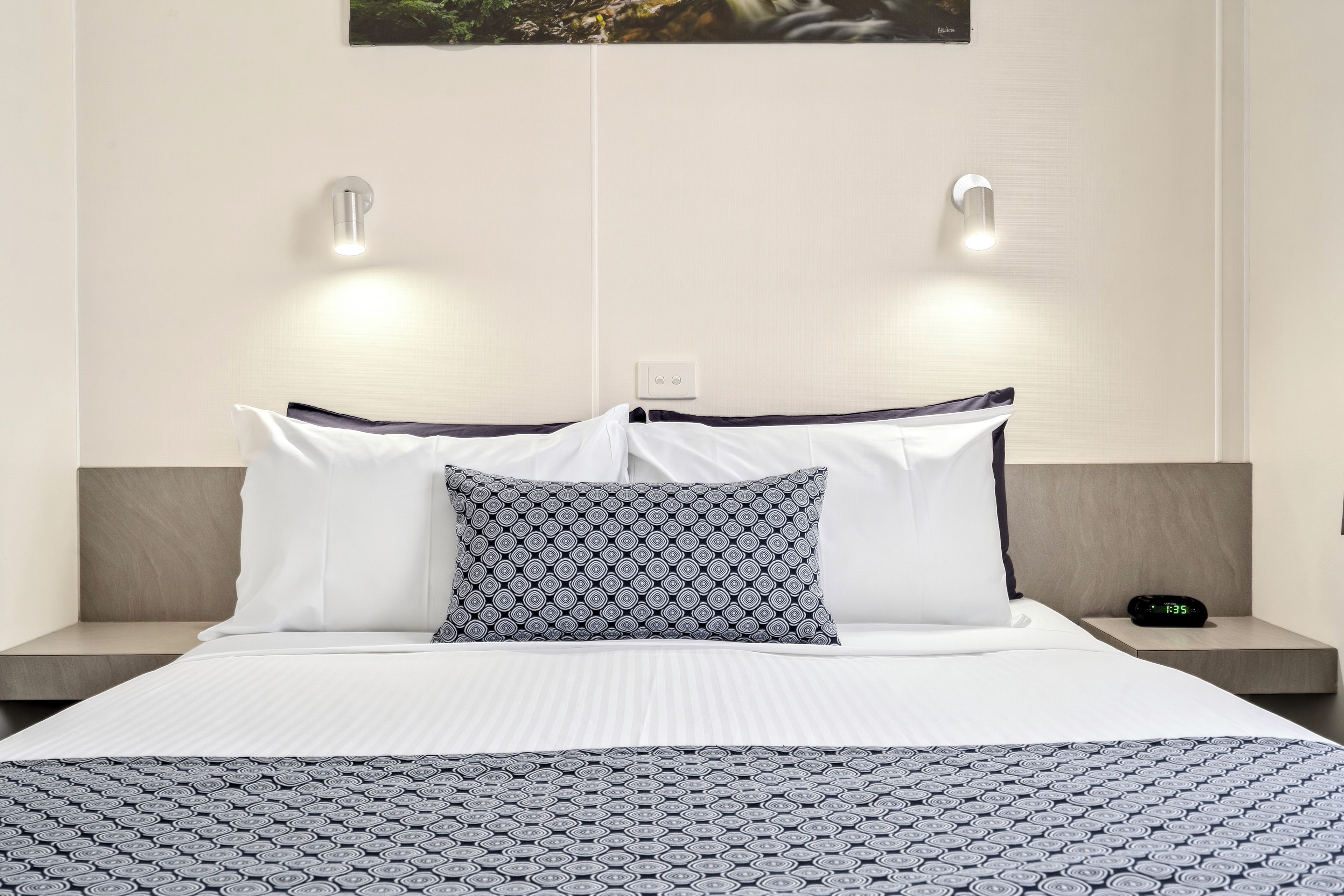 Una habitación de hotel con una cama, dos mesitas de noche, una lámpara de pared y un reloj digital.