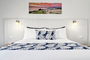 Una cama con ropa de cama blanca y azul, dos lámparas de pared y una pintura de paisaje en la parte superior.