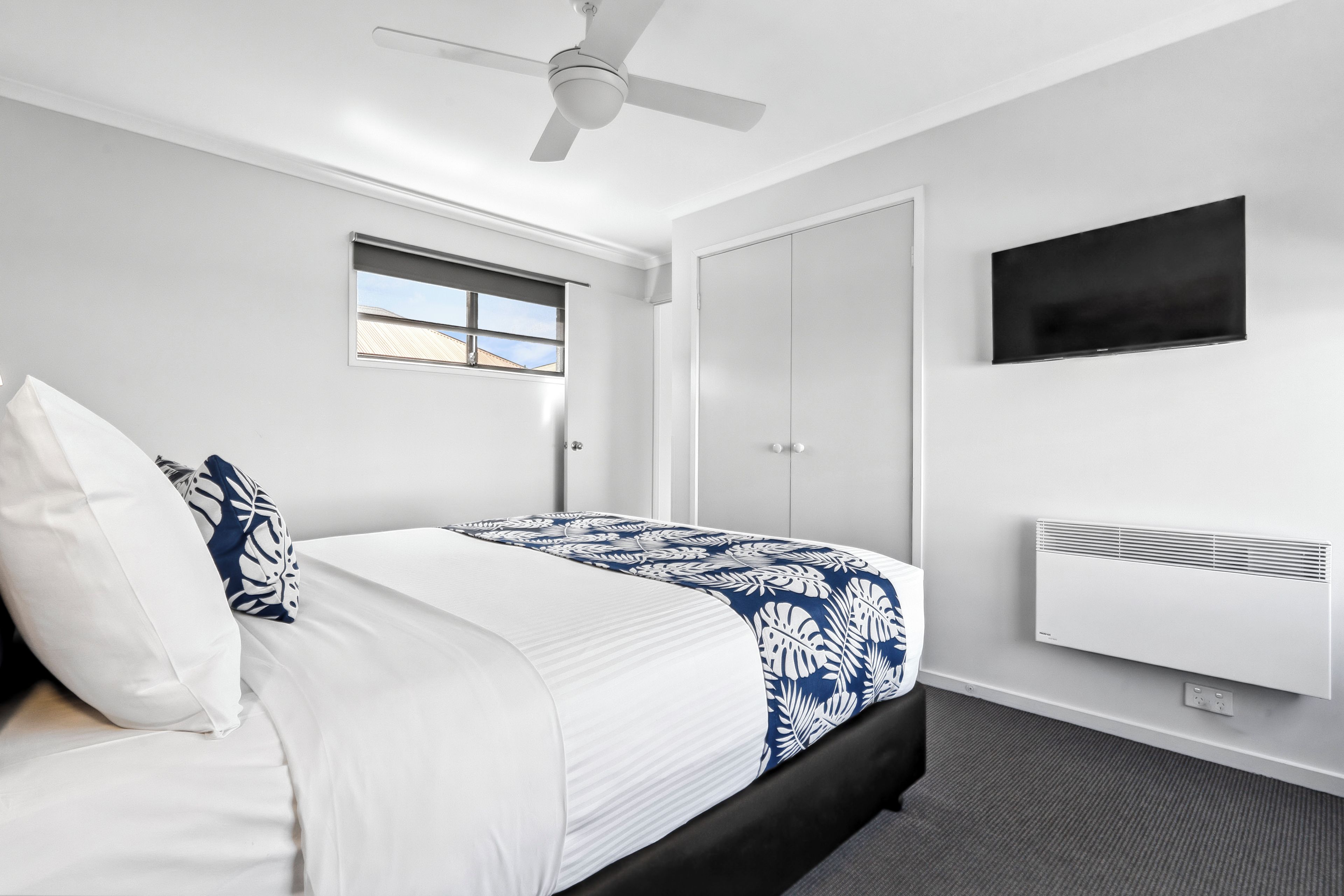 A bedroom with a bed, a ceiling fan, a window, a wardrobe, a flat-screen TV, and an air conditioning unit.