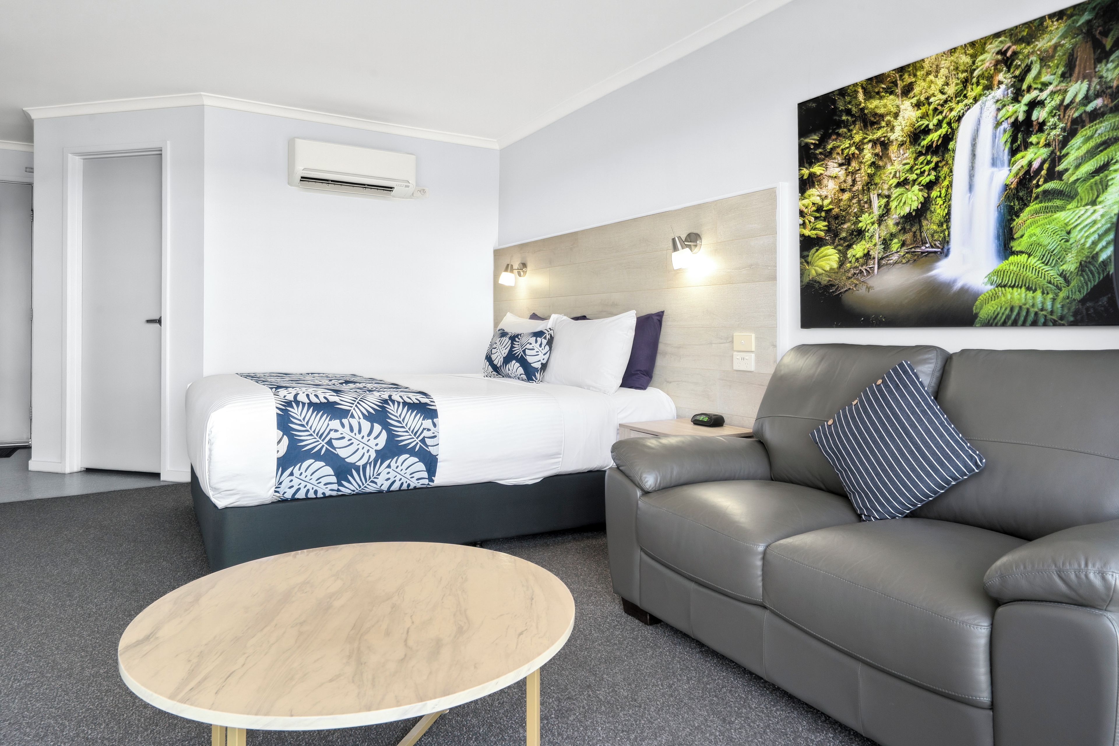 A modern hotel room with a bed, a grey sofa, a round wooden table, and a large wall-mounted picture of a waterfall.