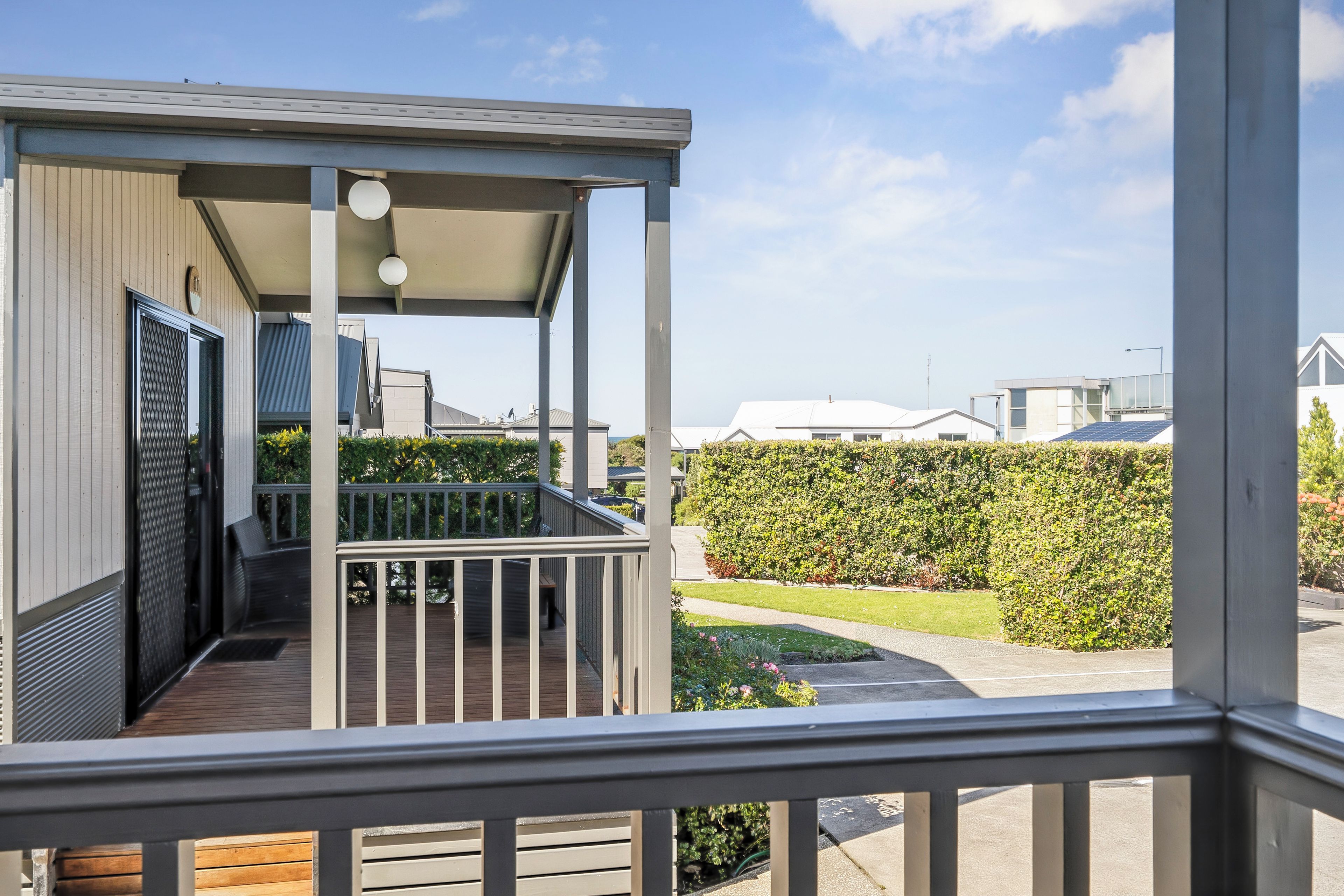 A covered outdoor area with a wooden deck, a metal railing, and a view of a neatly trimmed hedge and lawn.