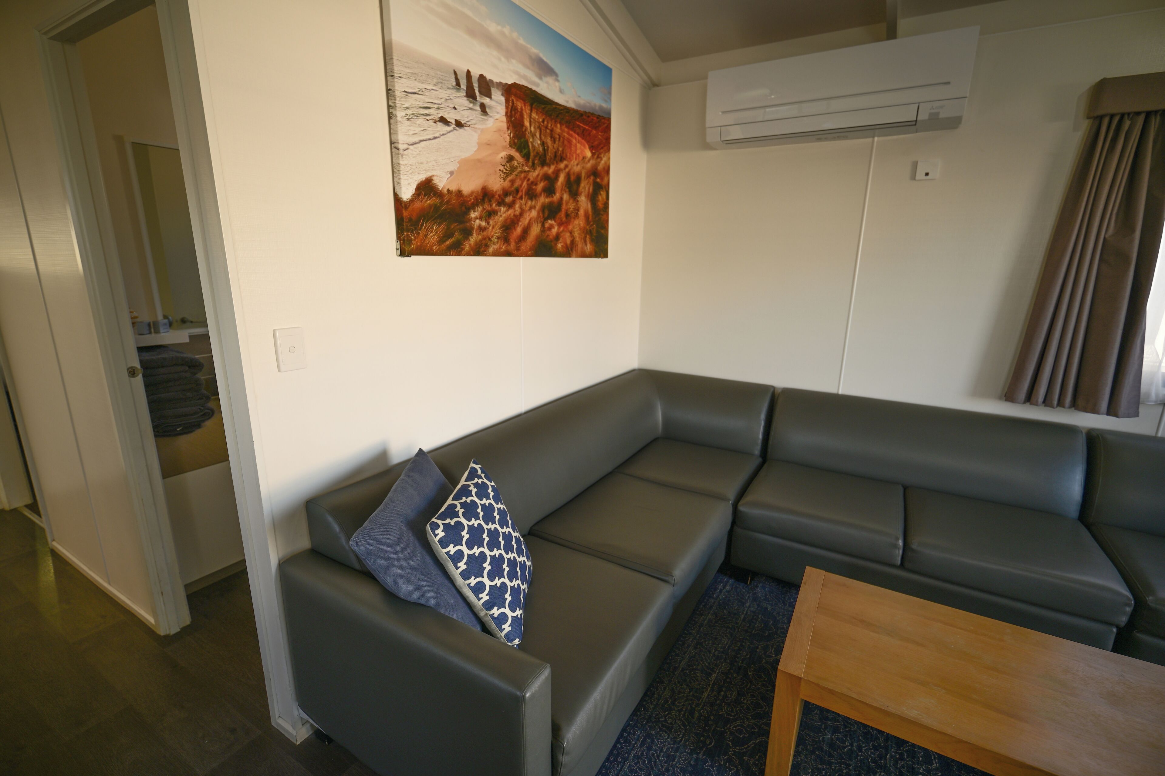 Een moderne woonkamer met een grijze modulaire bank, een decoratief kussen met een patroon, een houten salontafel en een schilderij van een kustlandschap aan de muur.