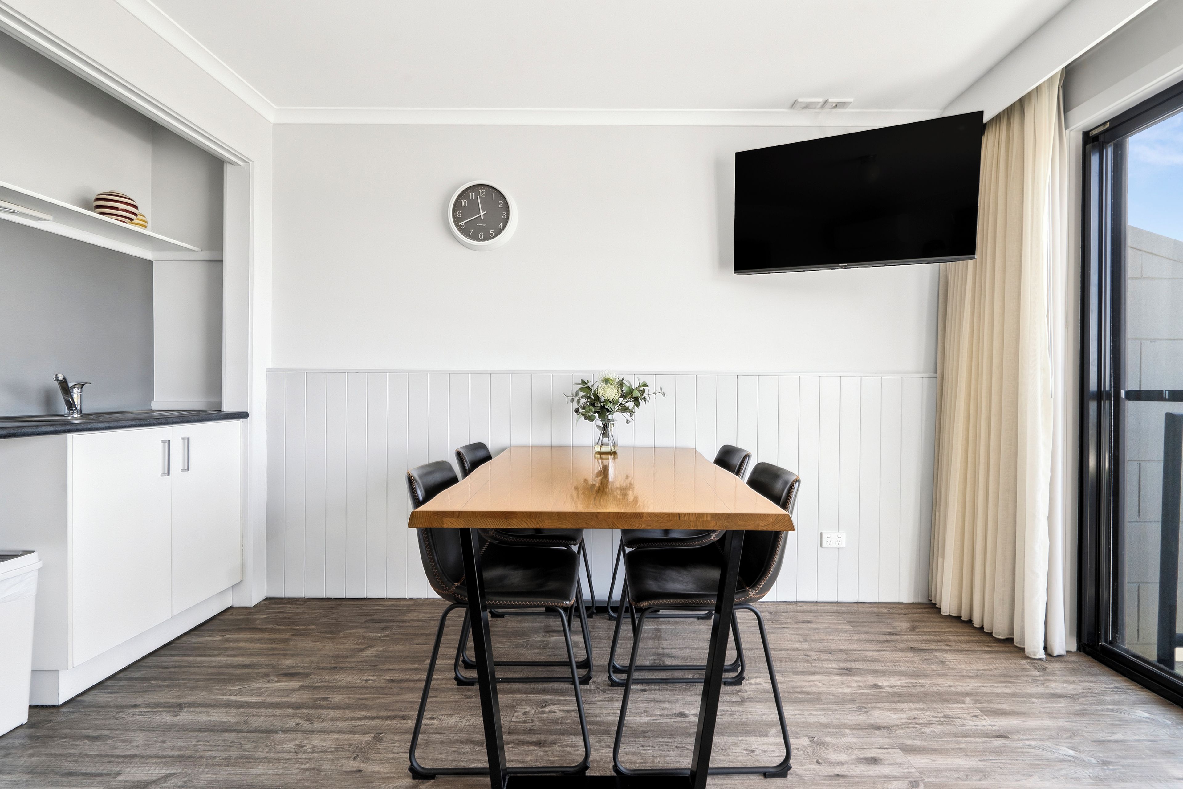 Una cocina moderna con armarios blancos, una mesa de madera con cuatro sillas, un reloj de pared y un televisor de pantalla plana.