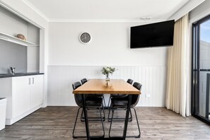 A modern kitchen with white cabinetry, a wooden dining table with four chairs, a wall-mounted clock, and a flat-screen TV.