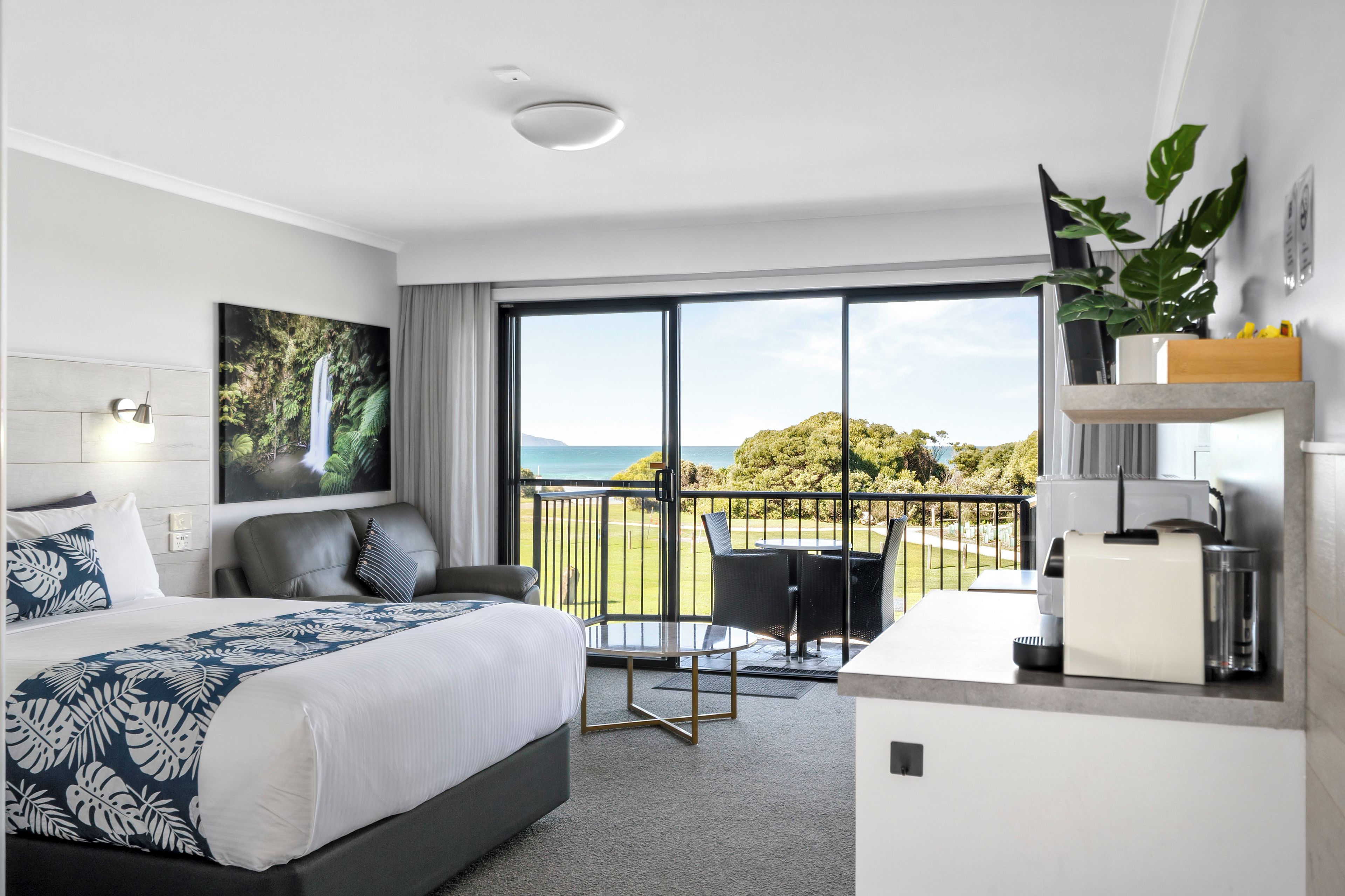 A hotel room with a bed, a desk, a chair, a balcony with a view of the sea, and a painting on the wall.