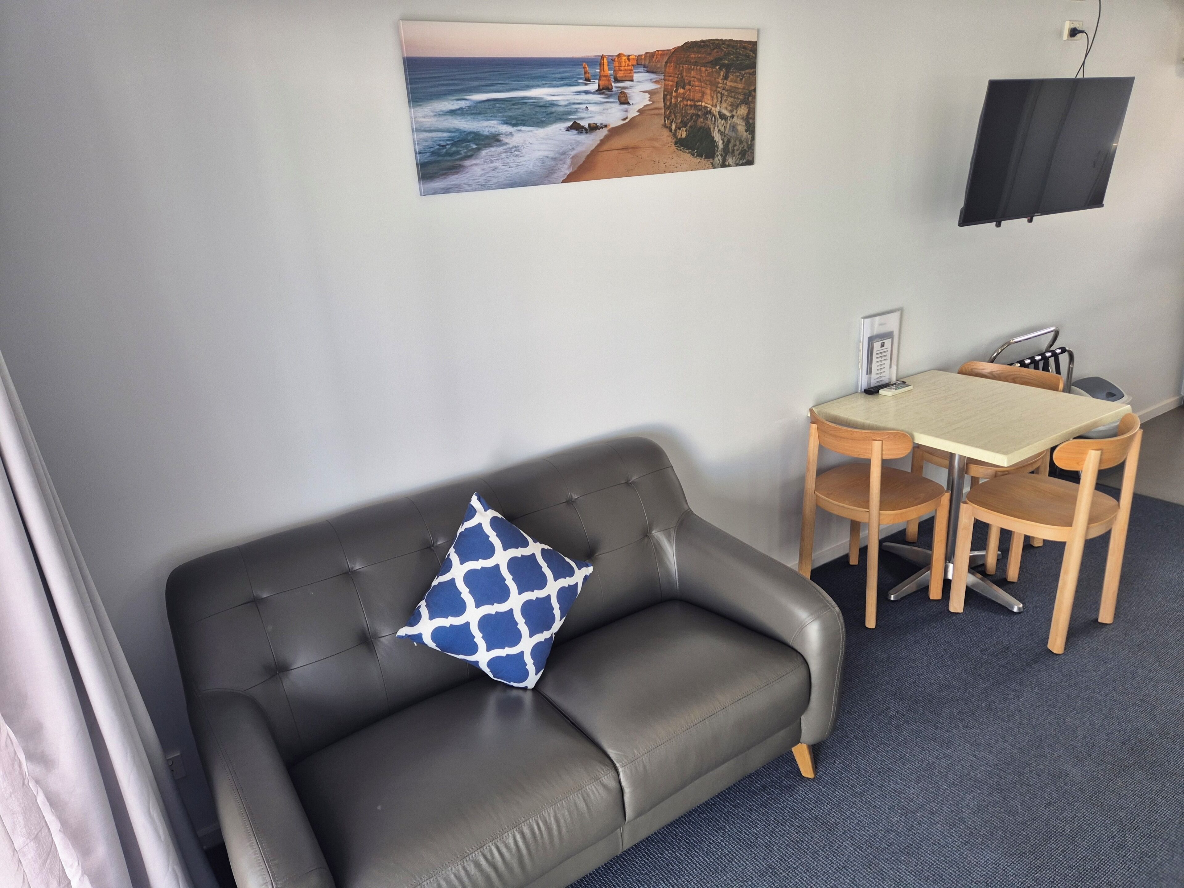 Una habitación con un sofá de cuero gris, una almohada con estampado azul y blanco, una mesita de madera con sillas, un televisor de pantalla plana montado en la pared y un cuadro con una escena costera.
