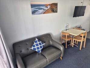 A room with a grey leather sofa, a blue and white patterned pillow, a small wooden table with chairs, a flat-screen TV mounted on the wall, and a framed picture of a coastal scene. - Apollo Bay Waterfront Motor Inn (Apollo Bay)