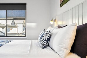 Une chambre d’hôtel avec un lit, une fenêtre donnant sur des immeubles et un luminaire fixé au mur.