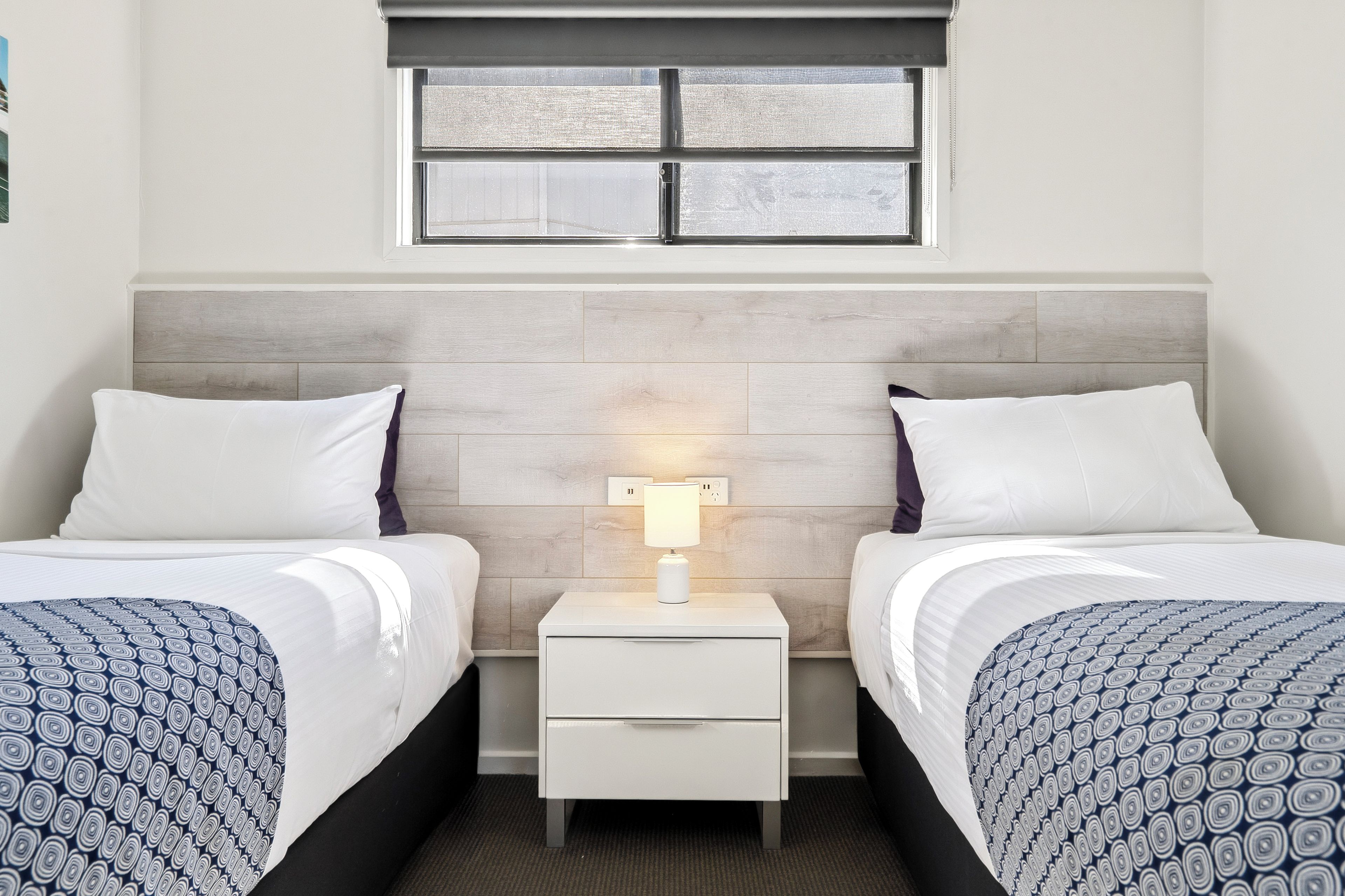 A hotel room with two beds, a nightstand with a lamp, and a window above the beds.