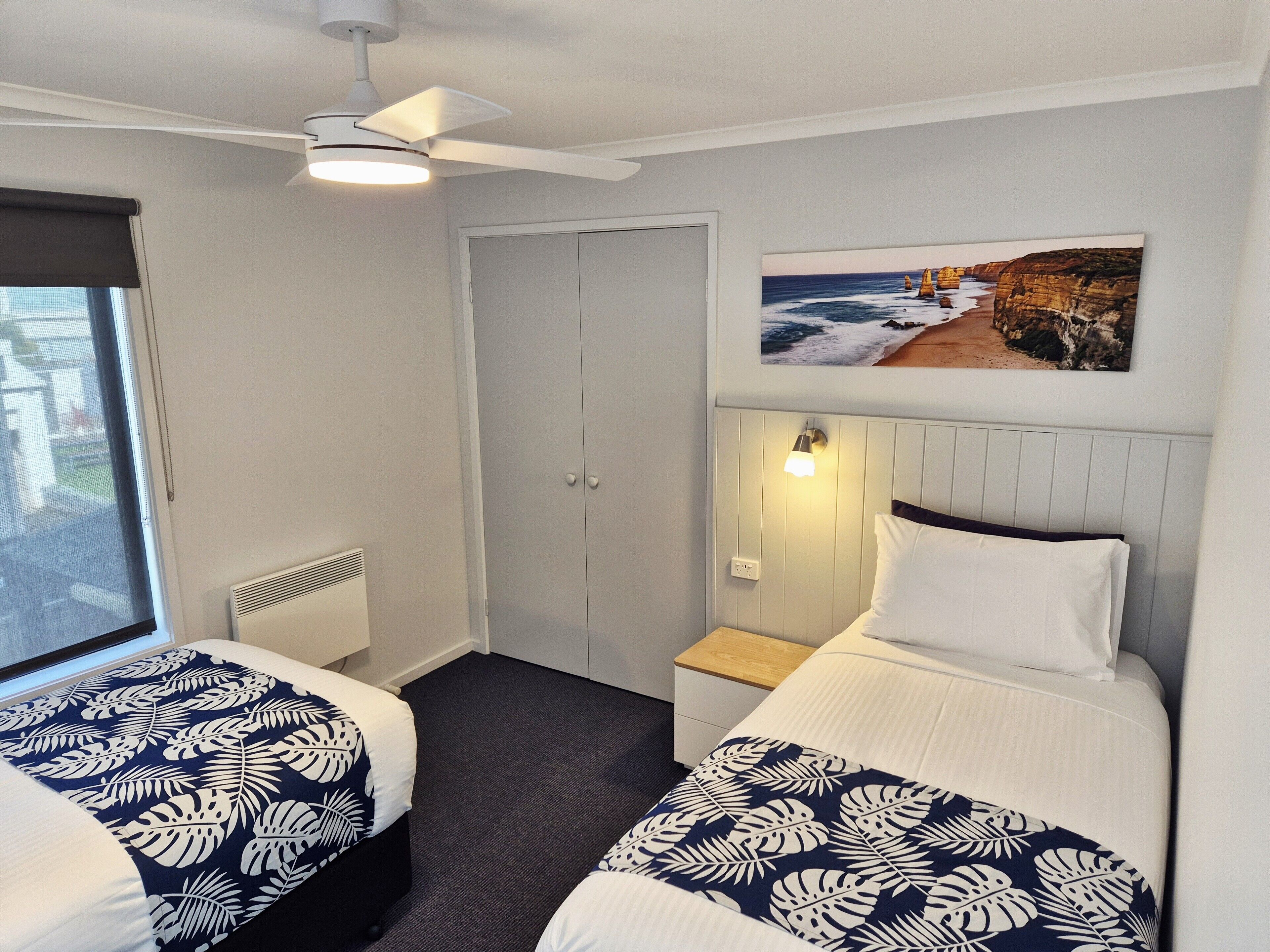 A hotel room with two beds, a ceiling fan, a painting of a coastal scene, and a window with a view.