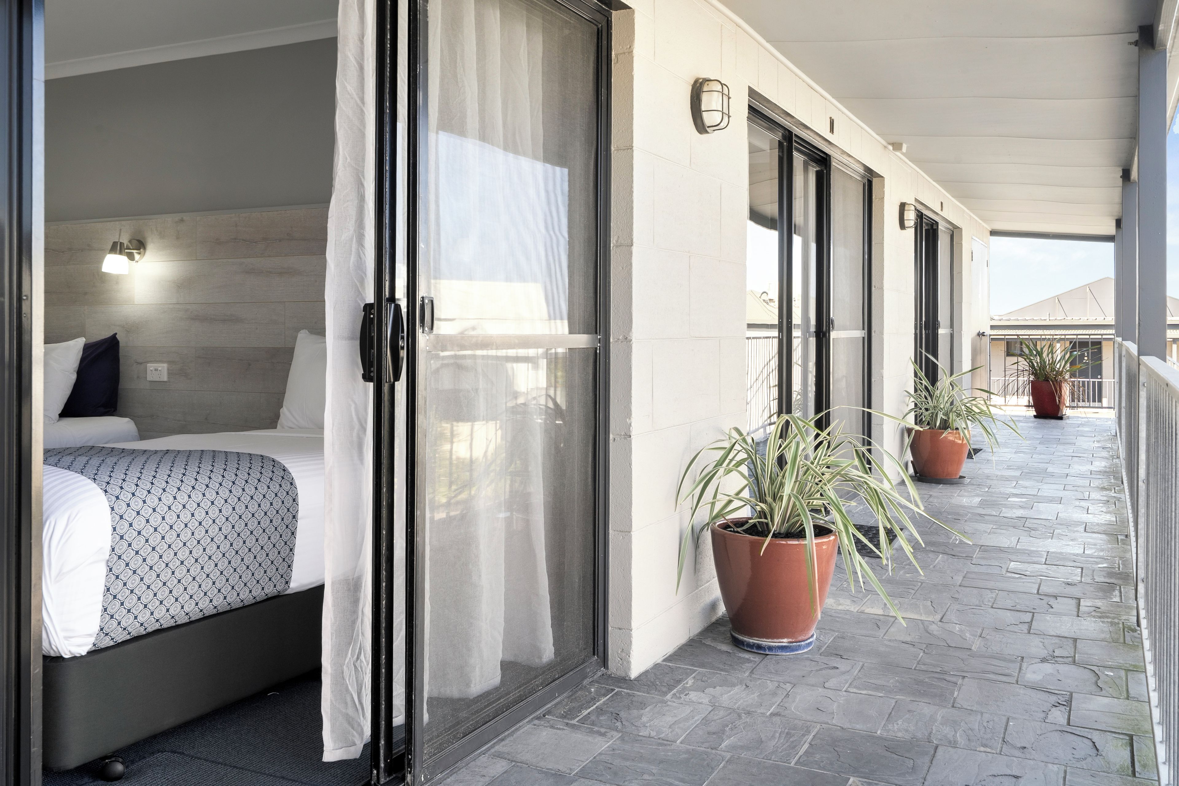 Habitación de hotel con cama, puerta de vidrio corrediza, planta en maceta y balcón con barandilla.