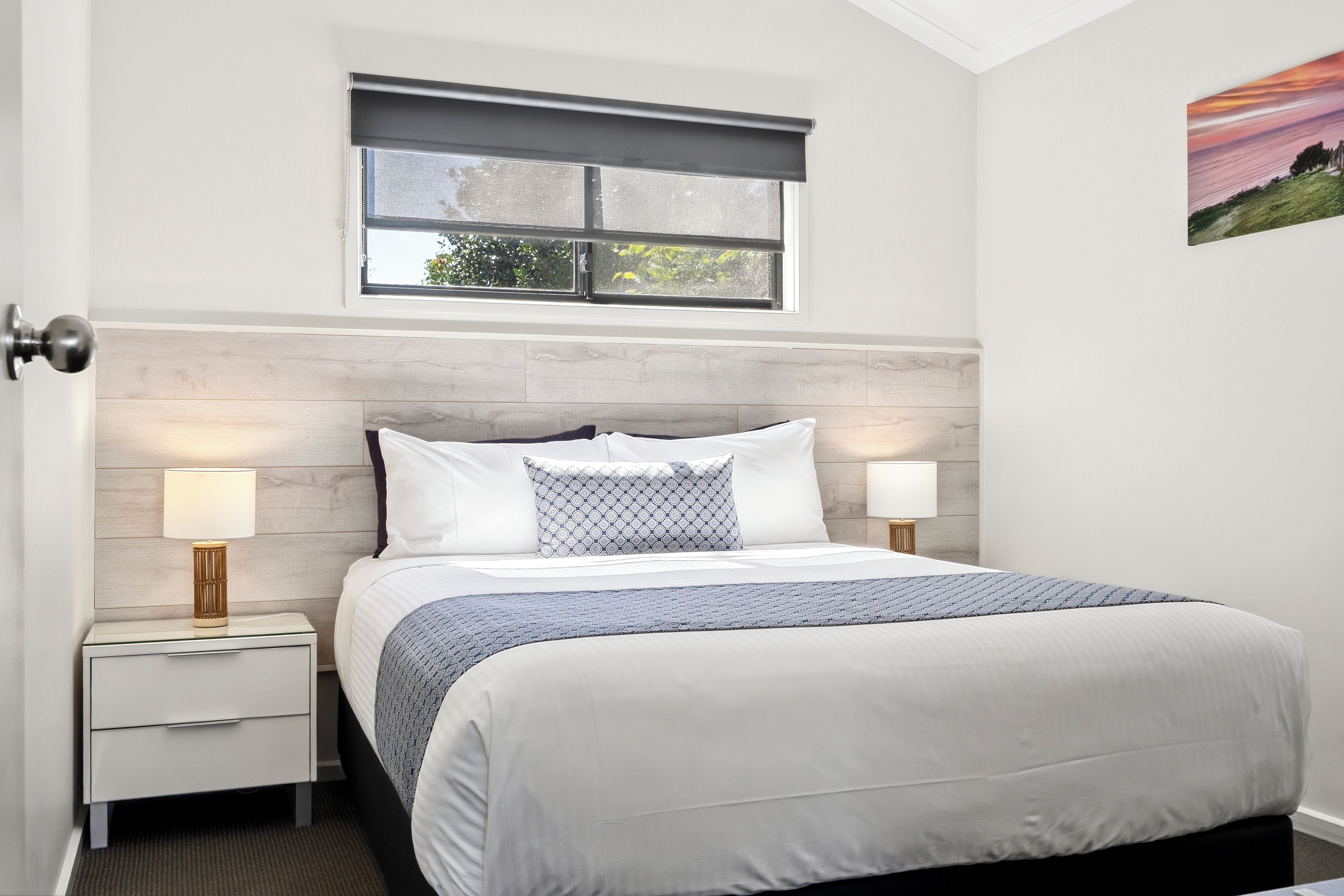 Quarto com cama, duas mesas de cabeceira com abajures, uma janela com cortina e um quadro na parede.