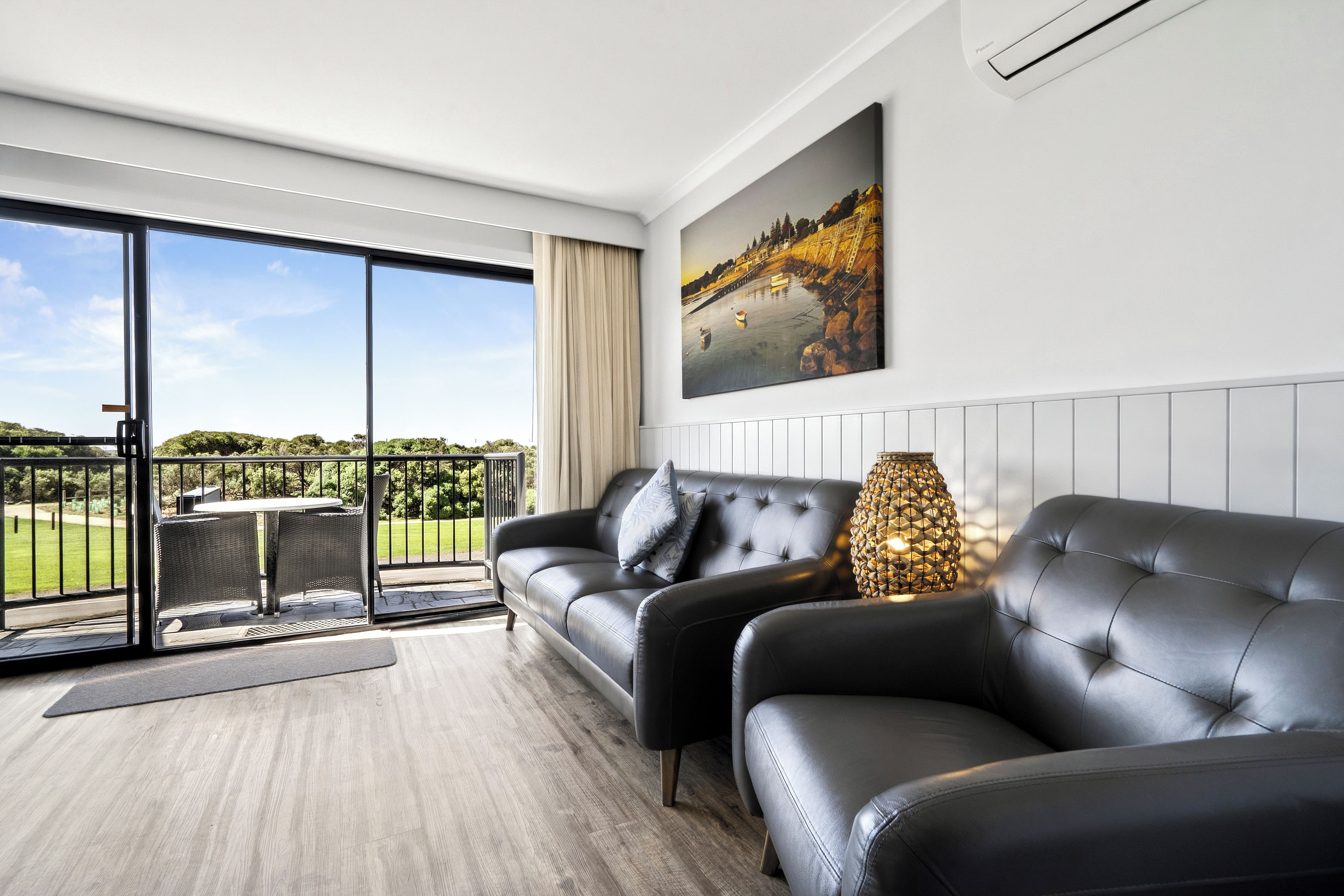 Ein modernes Wohnzimmer mit einem schwarzen Ledersofa, einem großen Fenster mit Blick auf eine grüne Landschaft und einem Gemälde einer Küstenansicht.