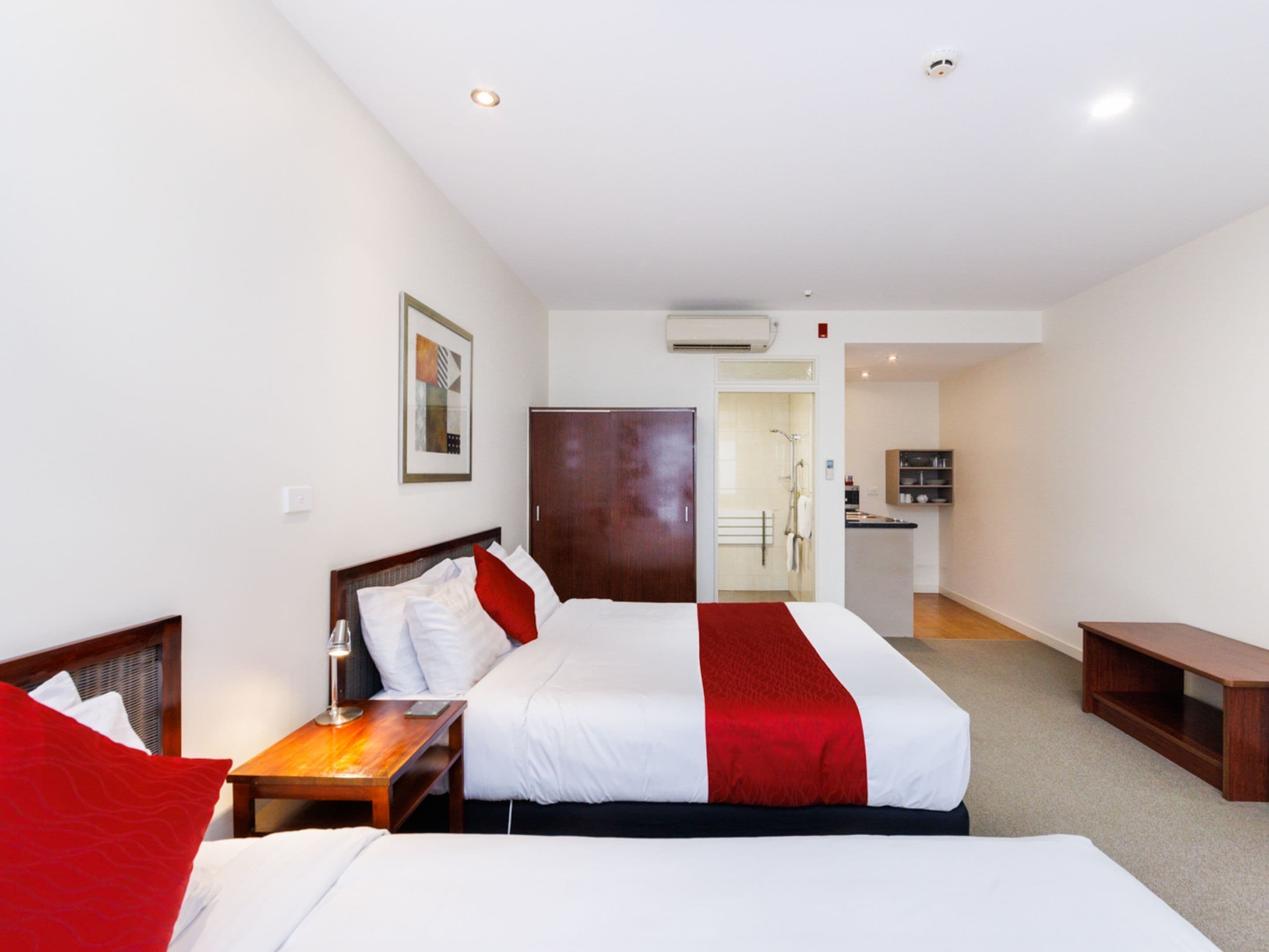 A hotel room with two beds, a wooden nightstand, a desk, a wardrobe, and a kitchenette in the background.
