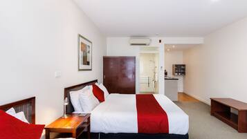 A hotel room with two beds, a wooden nightstand, a desk, a wardrobe, and a kitchenette in the background.