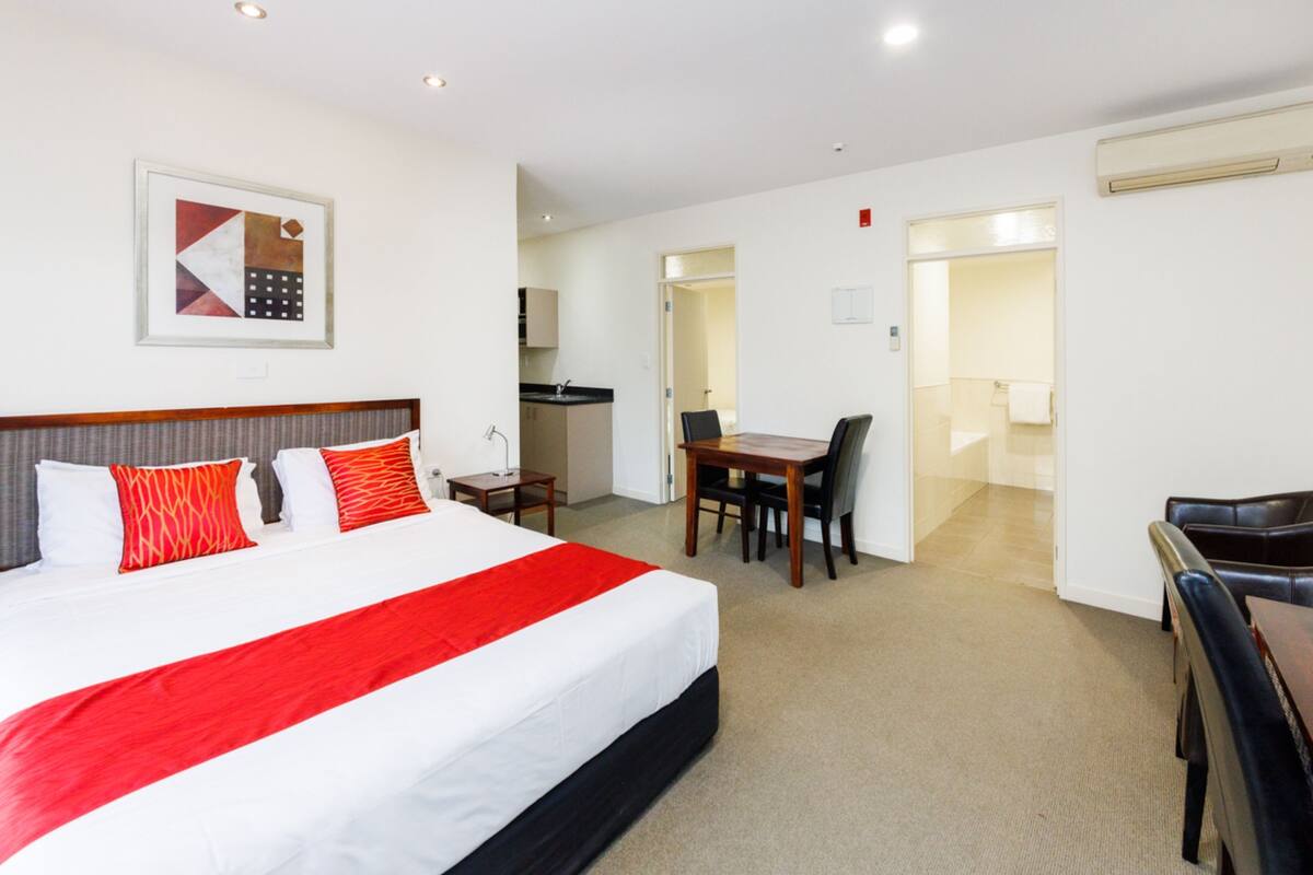 A hotel room with a large bed, a desk, and a chair. There is a kitchen area visible through an open door.