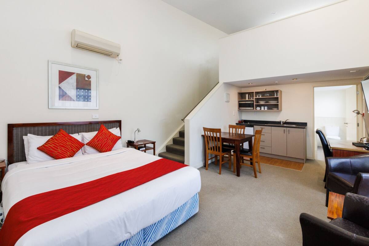 A bedroom with a bed, a desk, and a chair. There is a staircase leading to a kitchen area.