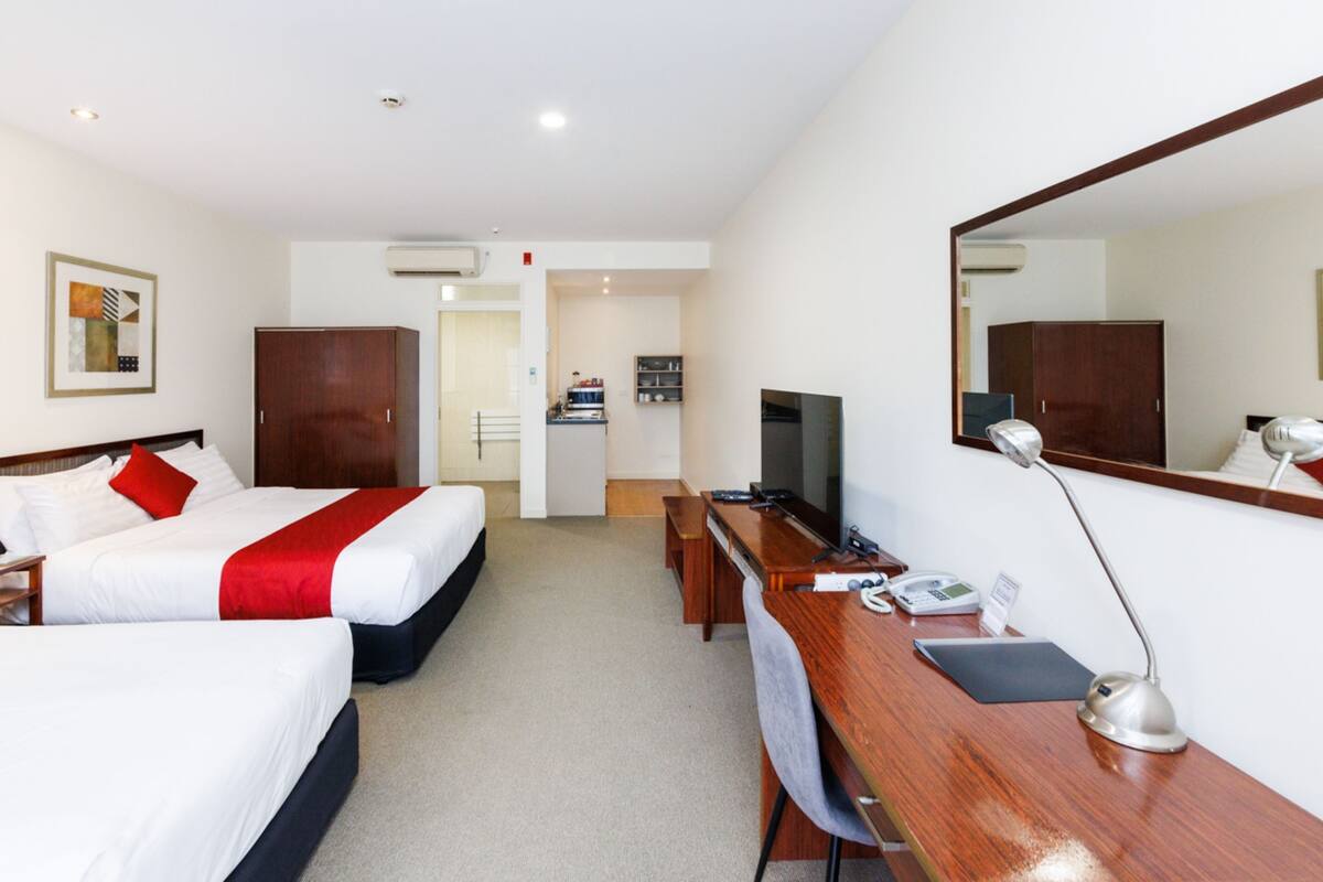 A hotel room with two beds, a desk, a TV, a mirror, and a kitchenette.