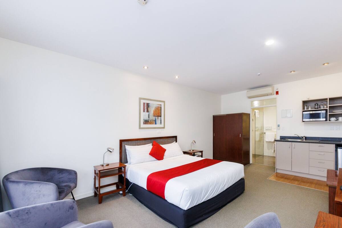 A hotel room with a bed, a desk, a chair, a wardrobe, and a kitchenette.