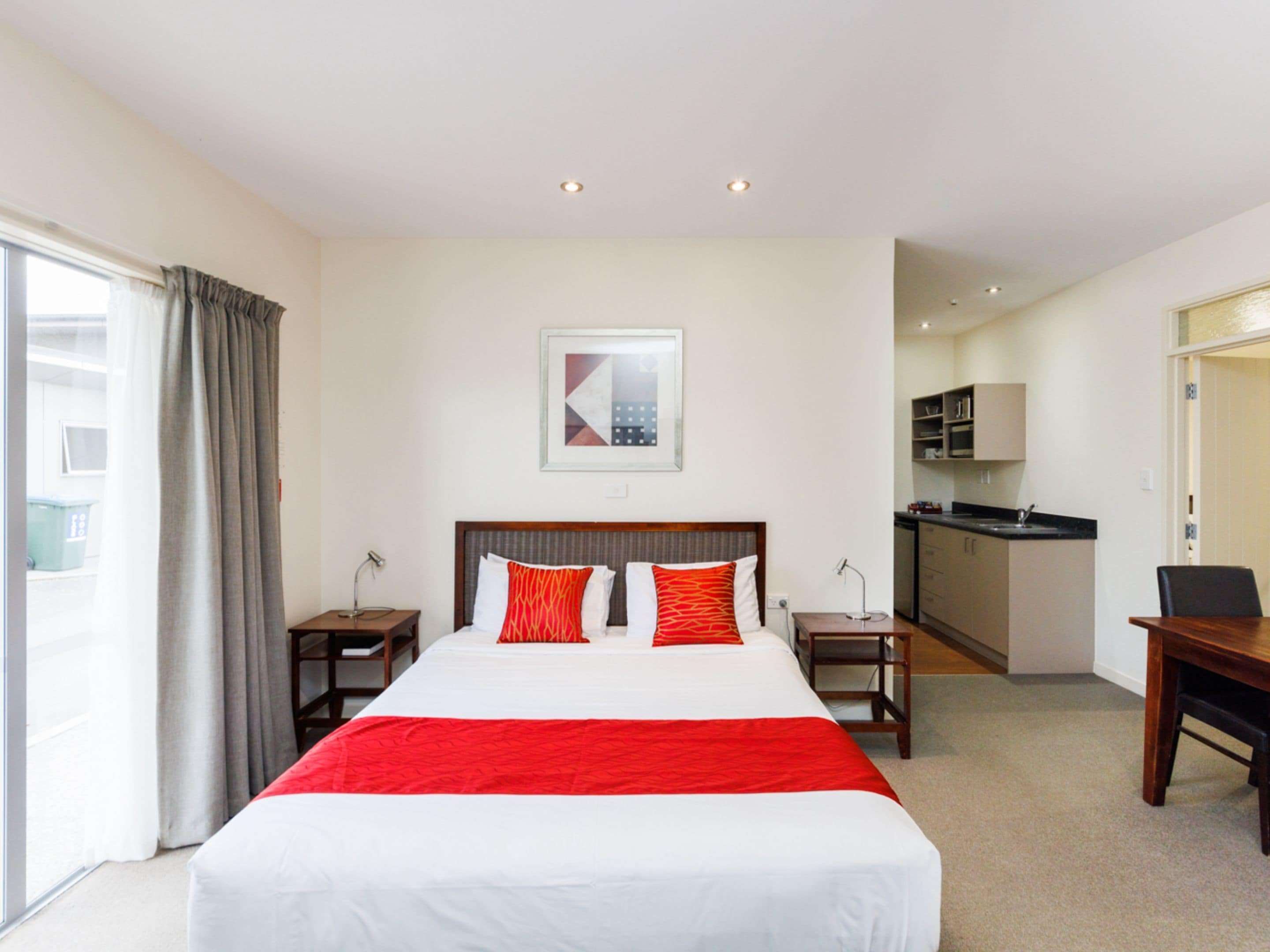 A hotel room with a large bed, a desk, a chair, a small table, a kitchenette, and a bathroom visible through an open door.
