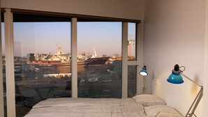 A bedroom with a large window, a bed, a desk, and a lamp.