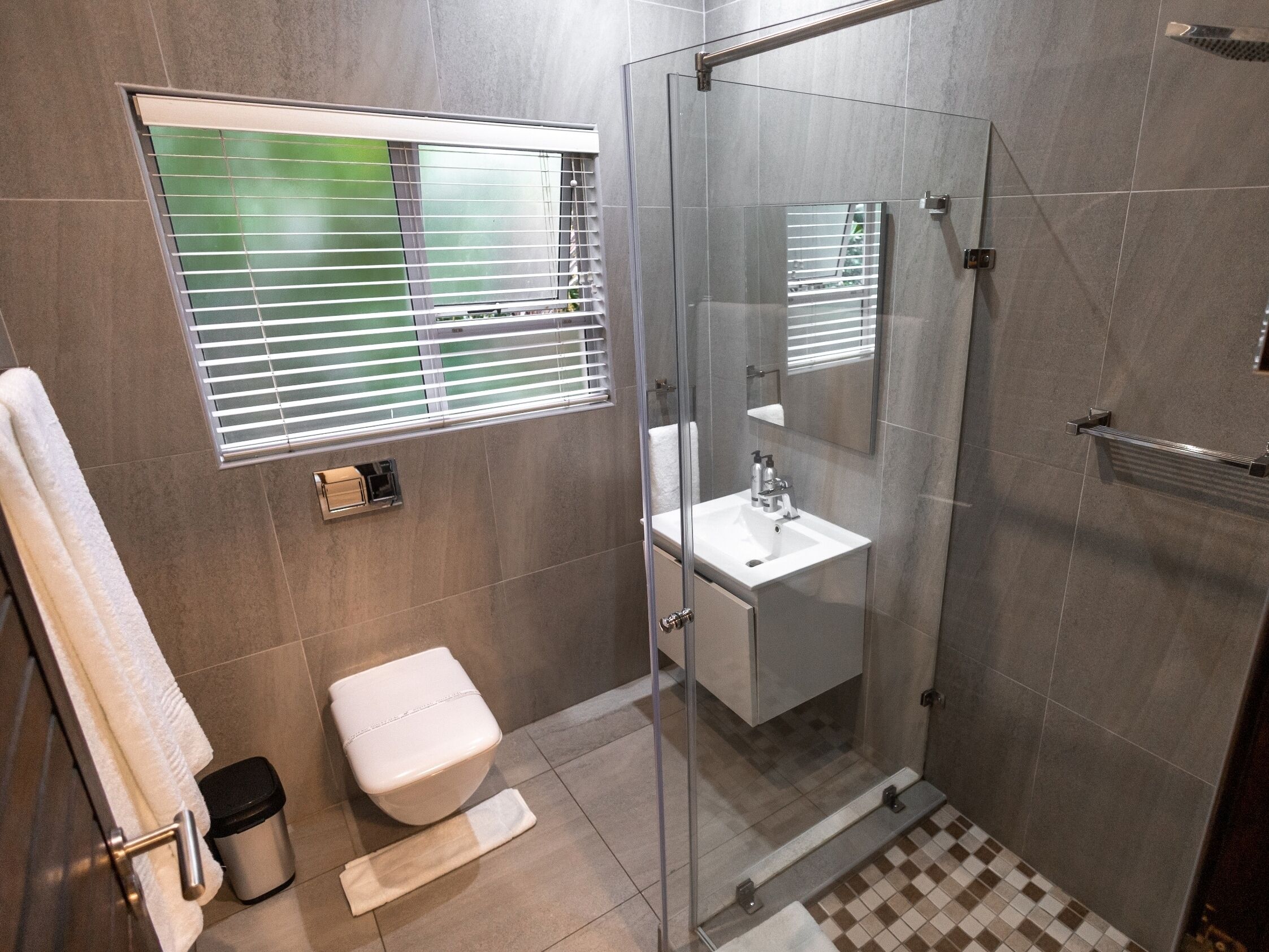 A modern bathroom with a glass shower enclosure, a toilet, a sink with a mirror, and a window with blinds.