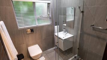 A modern bathroom with a glass shower enclosure, a toilet, a sink with a mirror, and a window with blinds.