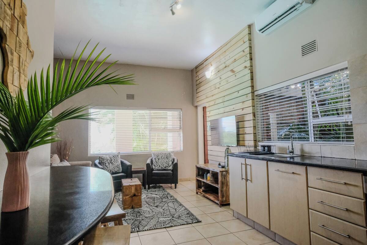 A modern kitchen with a dining area, featuring a large window, built-in cabinets, and a potted plant.