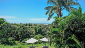 A lush outdoor area with two picnic tables and umbrellas, surrounded by dense tropical vegetation and a view of the ocean. - Little Eden St Lucia (St. Lucia)